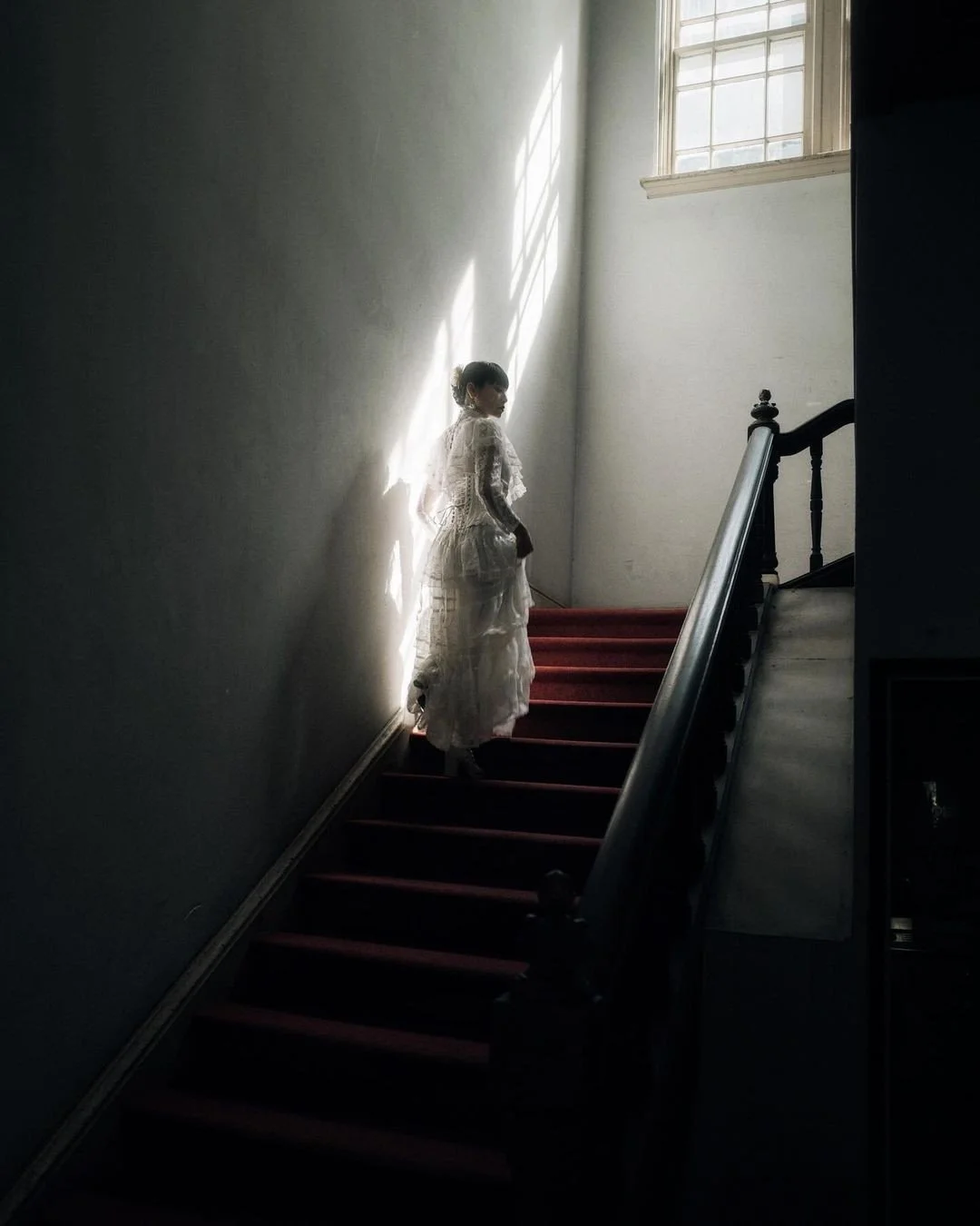 A woman in a vintage white dress stands on a staircase illuminated by sunlight coming through a window, with shadows cast on the wall.