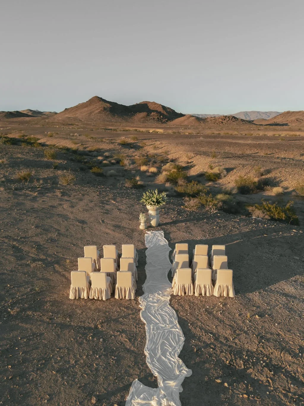 Outdoor desert scene with a wedding setup, including chairs with beige covers, a flowing white aisle runner, and two flower arrangements, with mountains in the background.