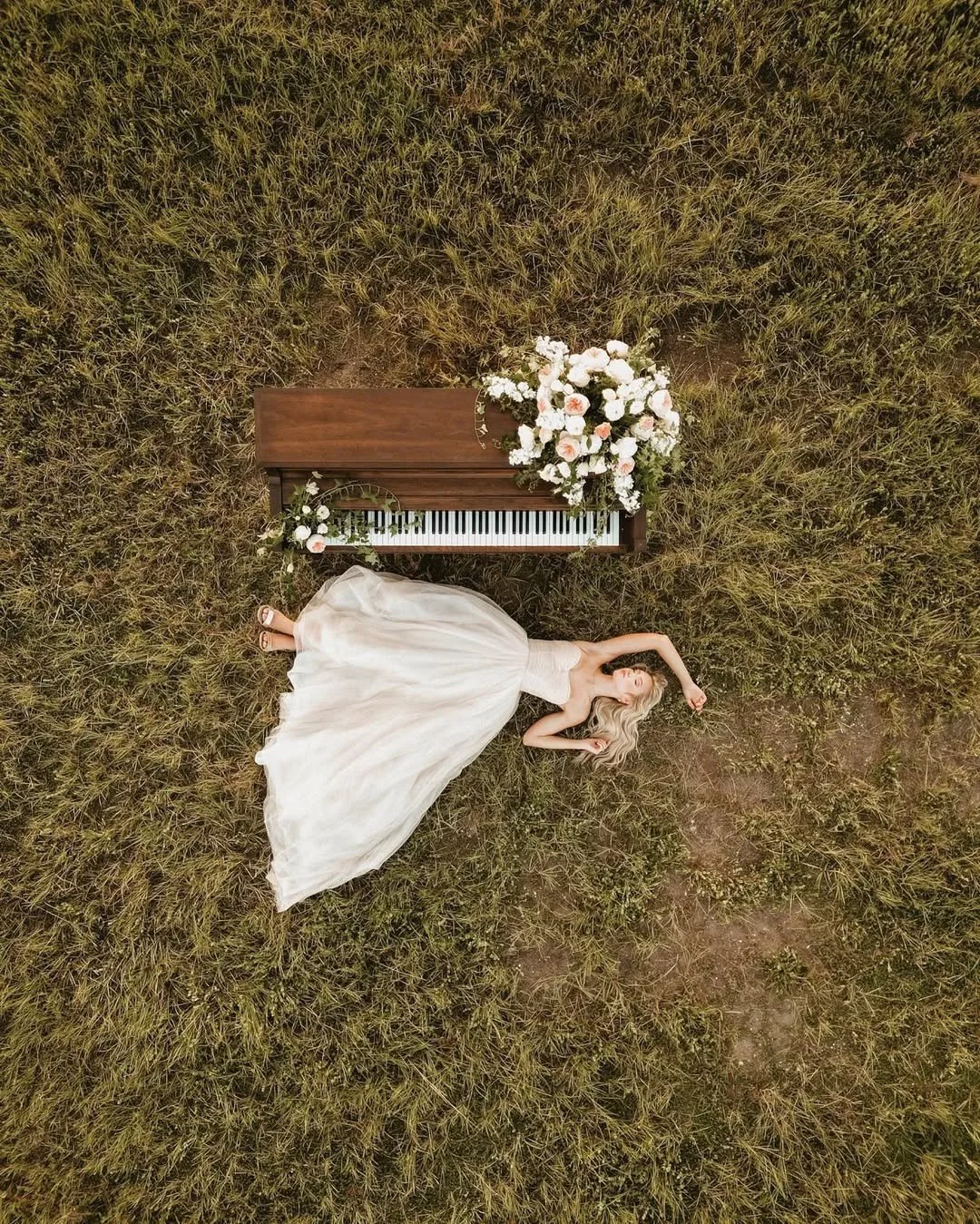 A woman in a white dress lies on the grass beside a wooden piano decorated with white and pink flowers.