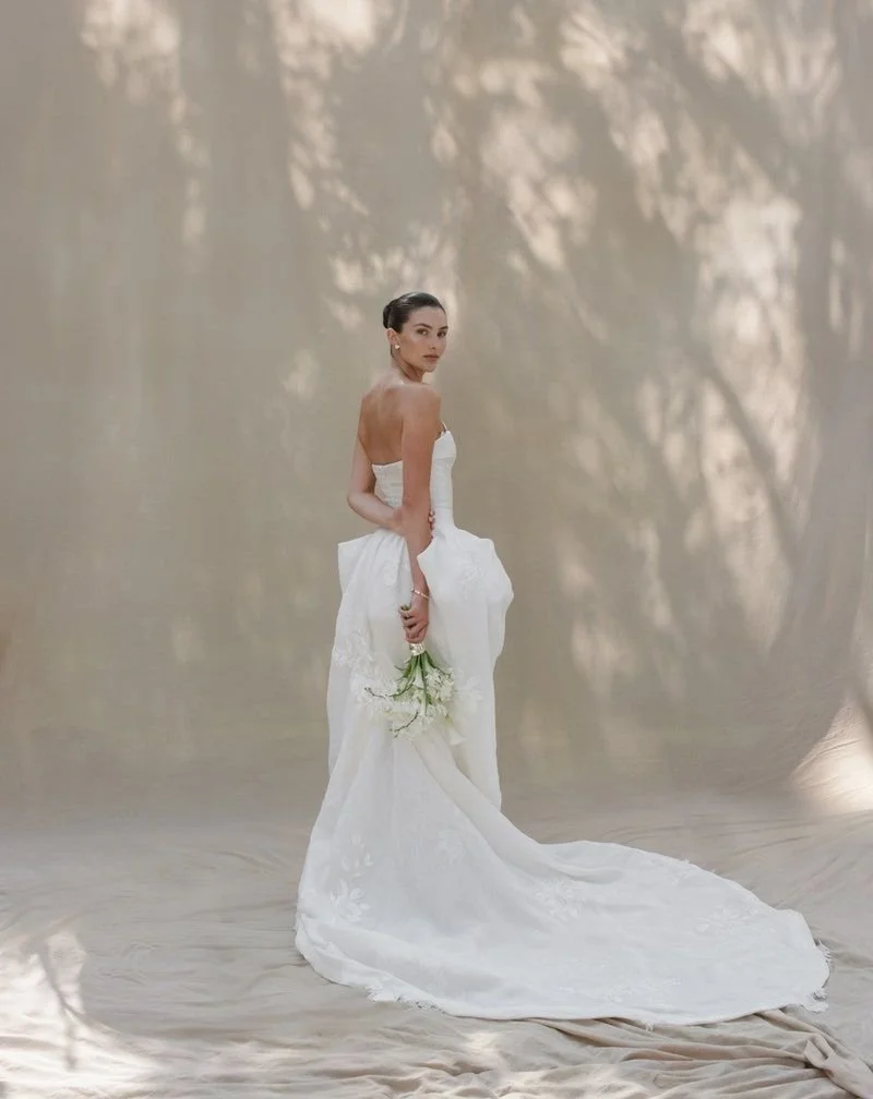 A bride in a strapless white wedding gown holding a small bouquet of white flowers, standing on a neutral backdrop with shadows of trees.