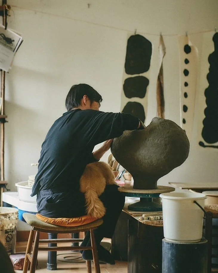 A person working on a large clay sculpture inside a studio, with a dog sitting on a cushion on their lap.