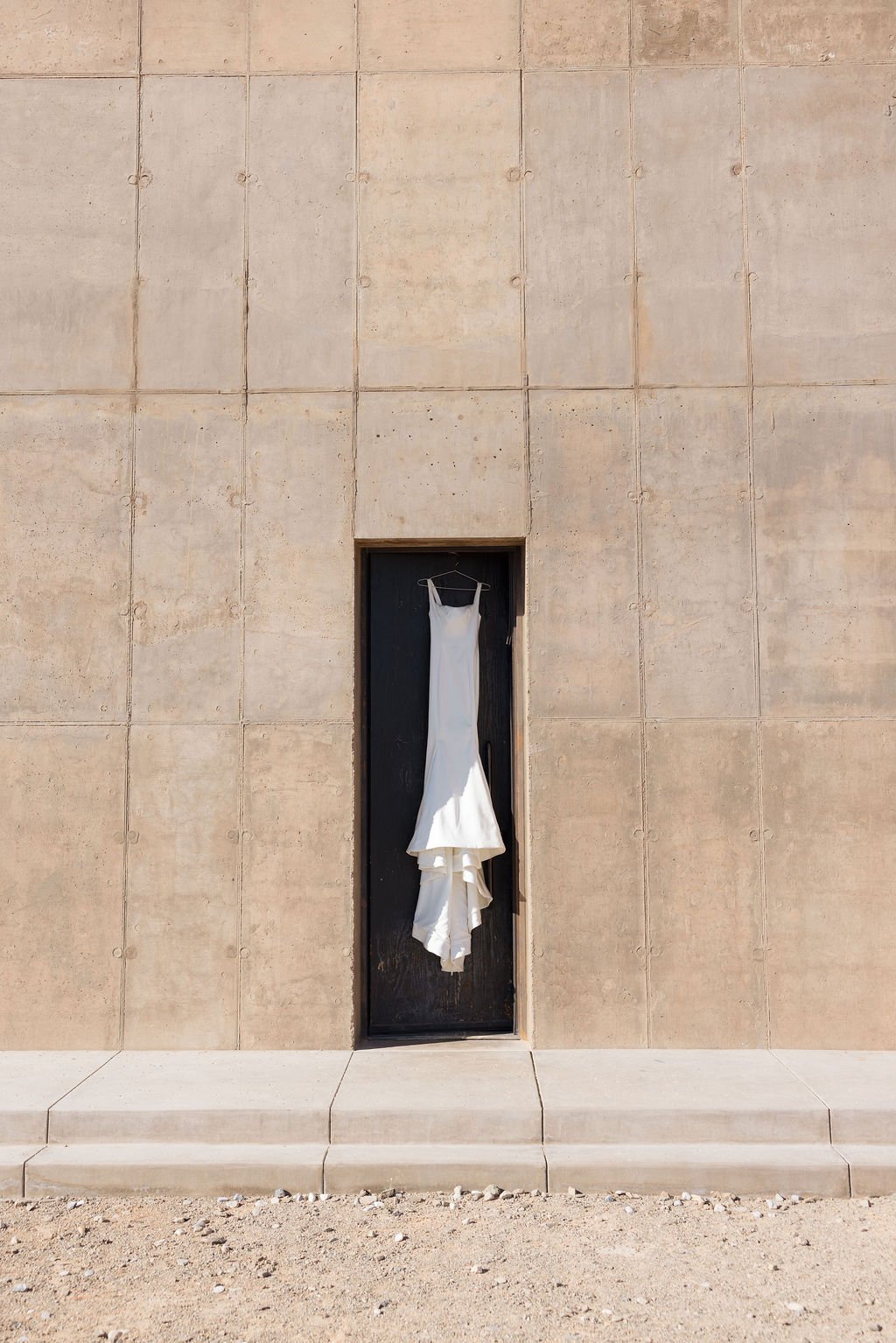 A white wedding dress hanging on a hanger inside a narrow black doorway on a beige stone wall.