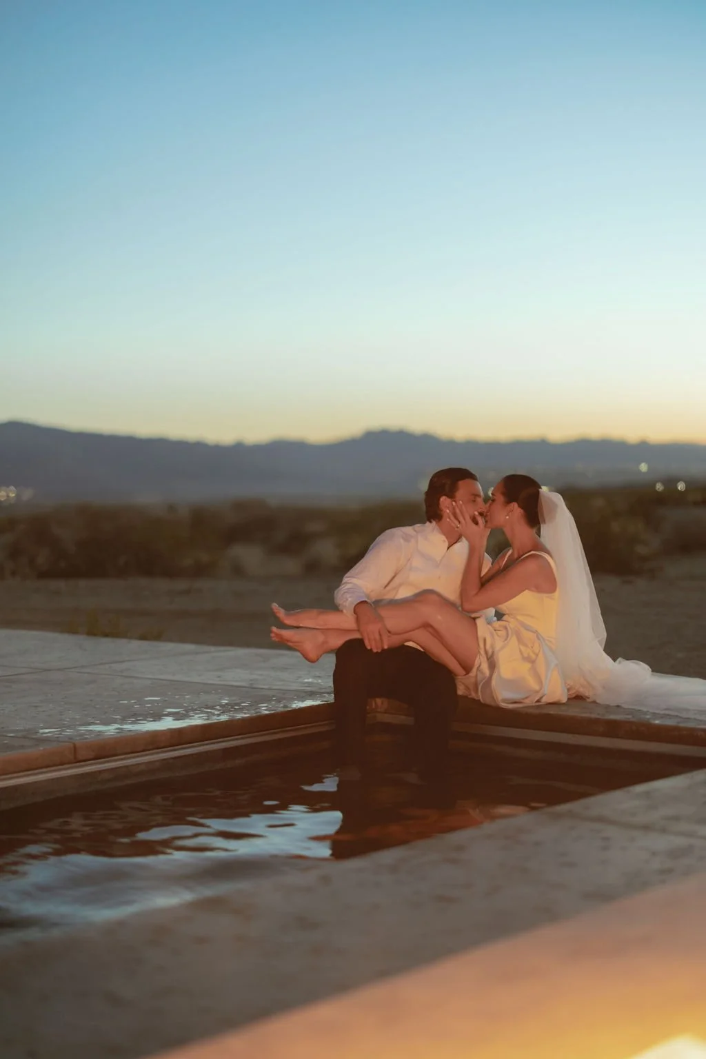 A couple in wedding attire sharing a kiss while sitting on the edge of a pool during sunset with mountains in the background.