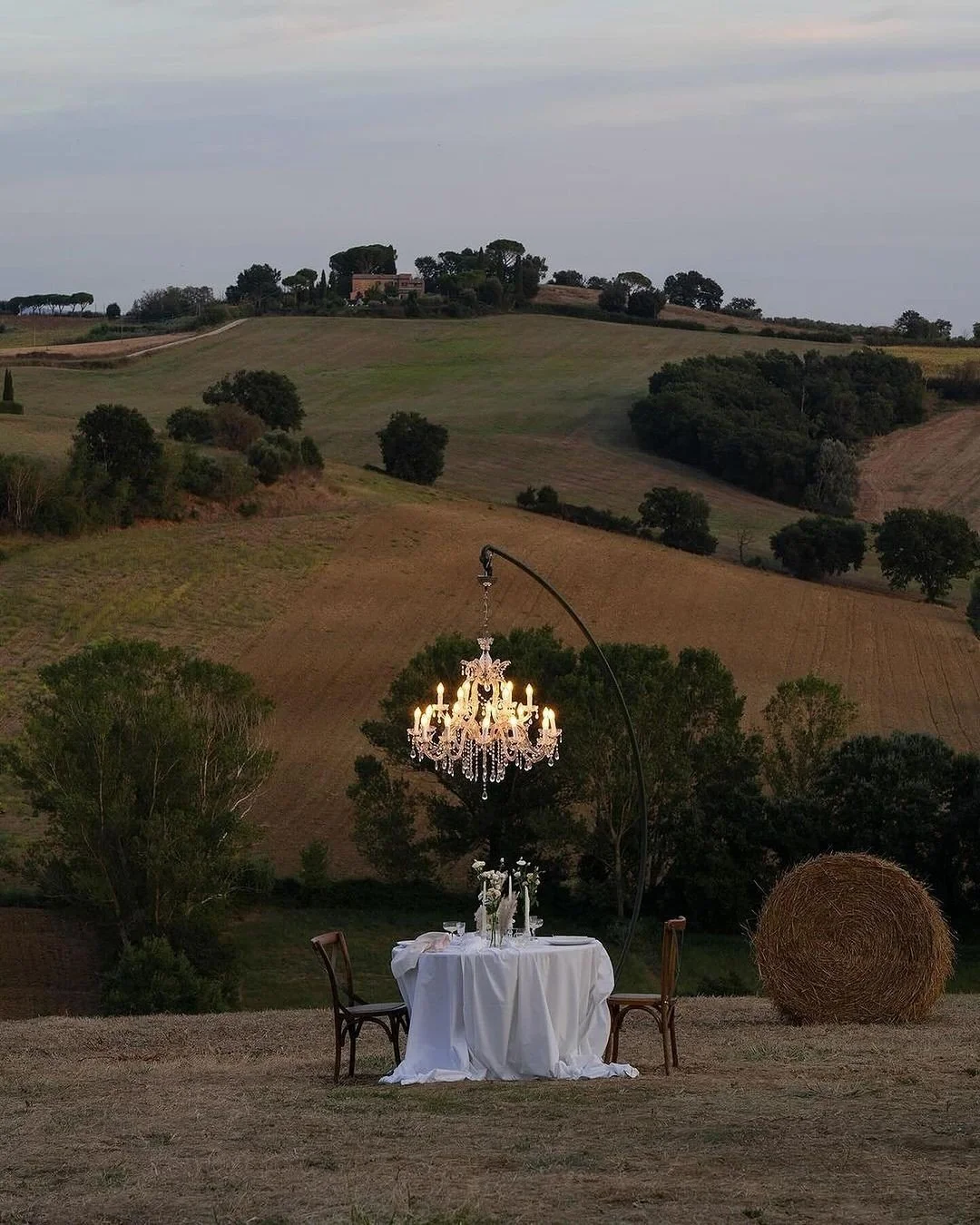 A table with a white tablecloth, a crystal chandelier hanging overhead, two wooden chairs, and a hay bale on a grassy hillside with rolling green hills and trees in the background.