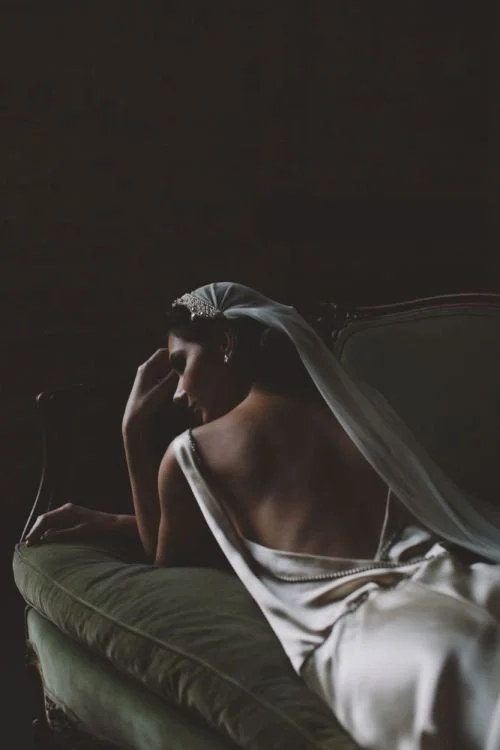 A woman in a wedding dress with a veil, sitting on a vintage-style bed in a dimly lit room, gazing downward with her hand near her face.