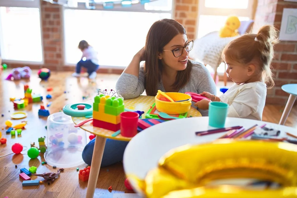Female child care teacher sitting at an activity table and talking with a young girl.