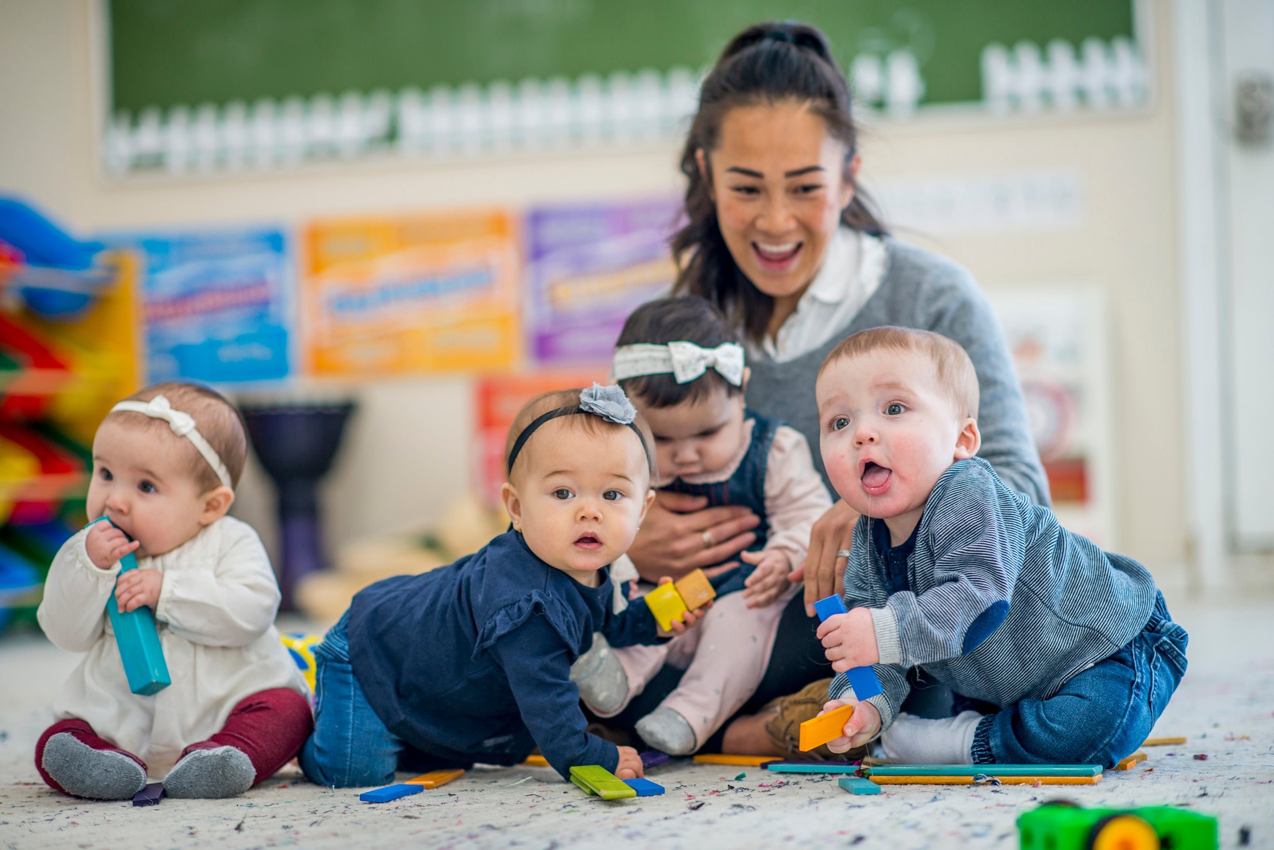 Female teacher in a gray sweater sitting on the floor of a child care room with four babies, one of which is on her lap.