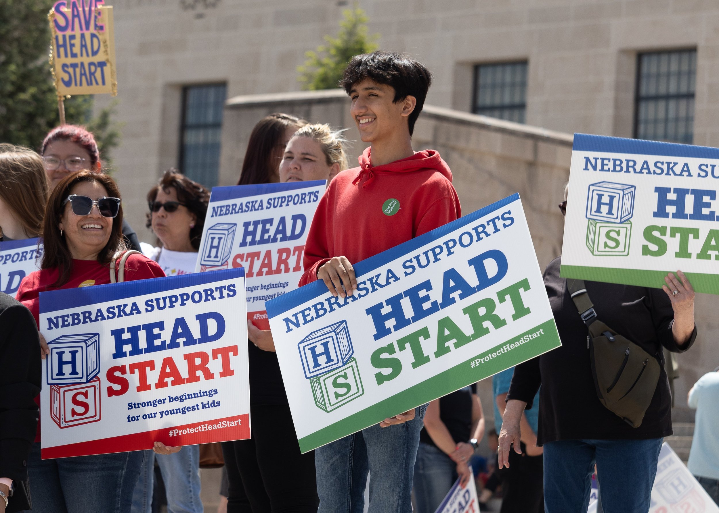 Man in a red shirt attending an outdoor rally and holding a sign that reads "Nebraska Supports Head Start"