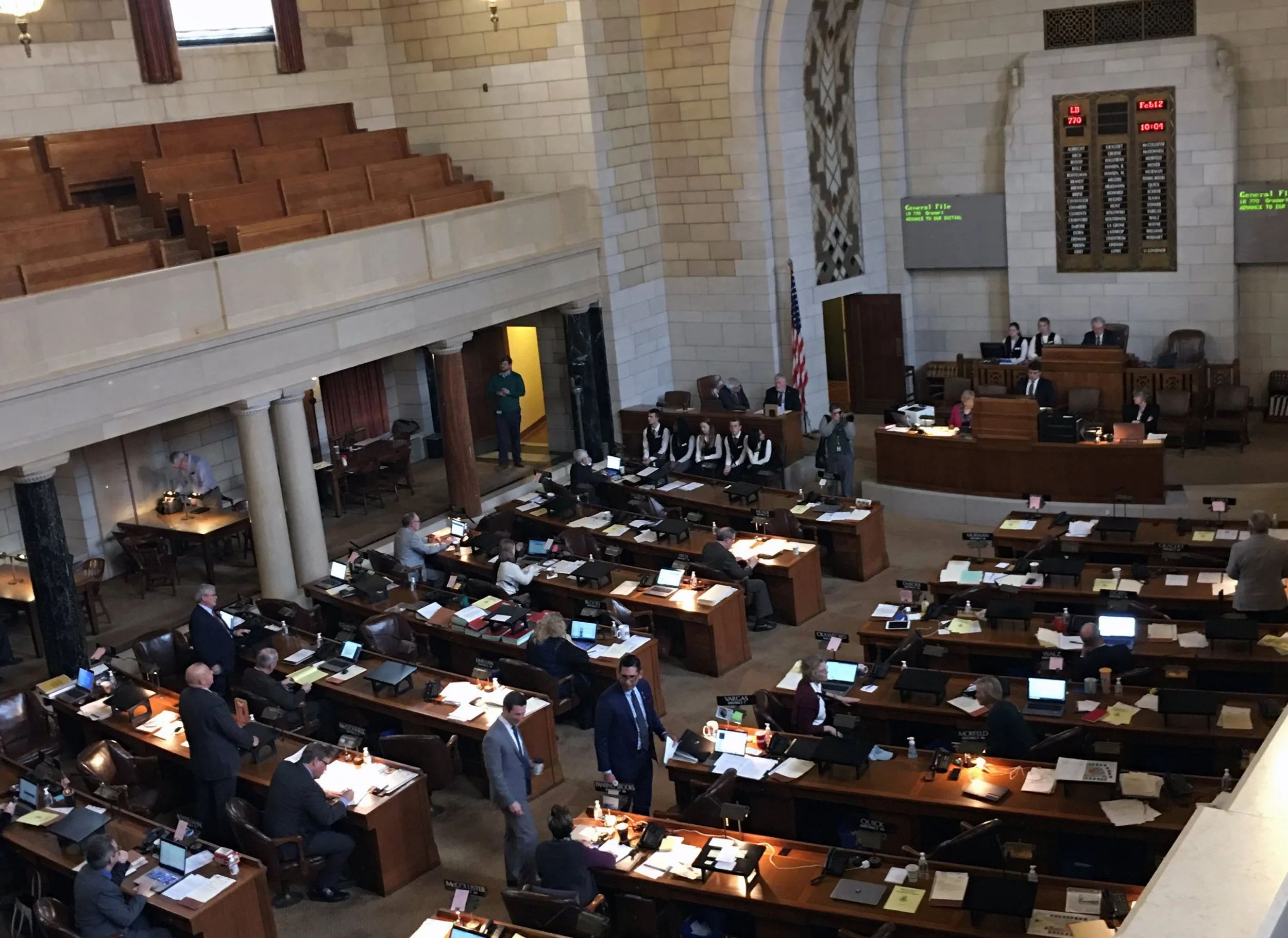 Overhead view of senators standing around wooden desks in the Nebraska legislative chamber..