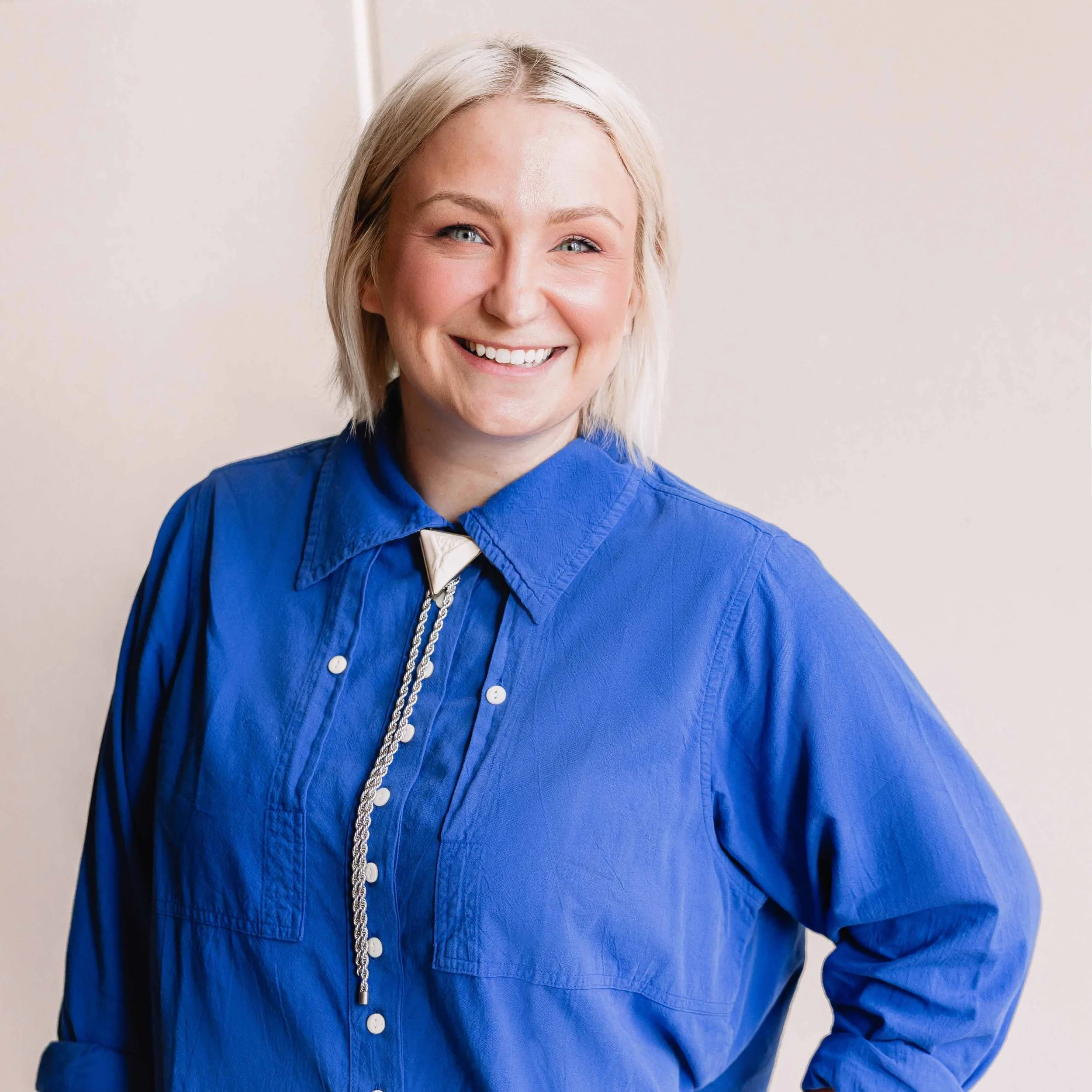 A smiling woman with short blonde hair wearing a royal blue button-up shirt and a silver necklace, standing against a plain light-colored background.