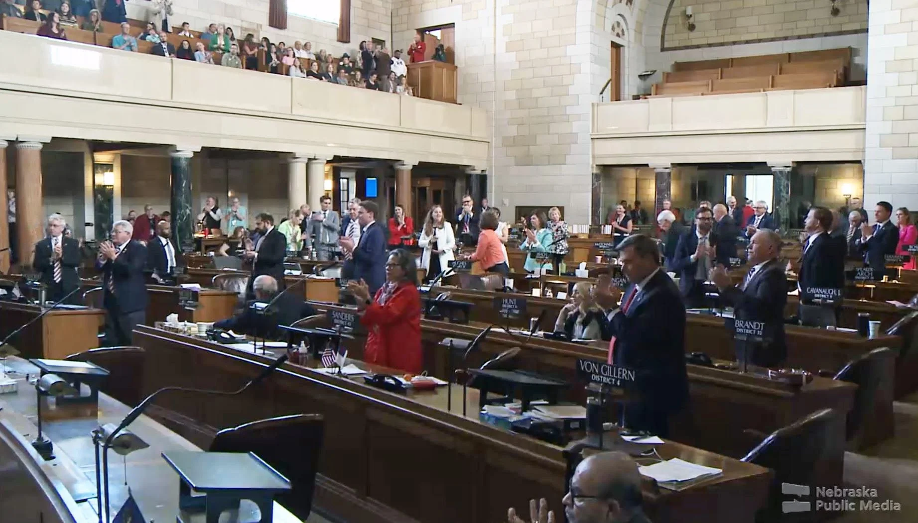 Crowd of people standing at wooden desks and on a balcony overlooking the legislative chamber of the Nebraska Capitol building.