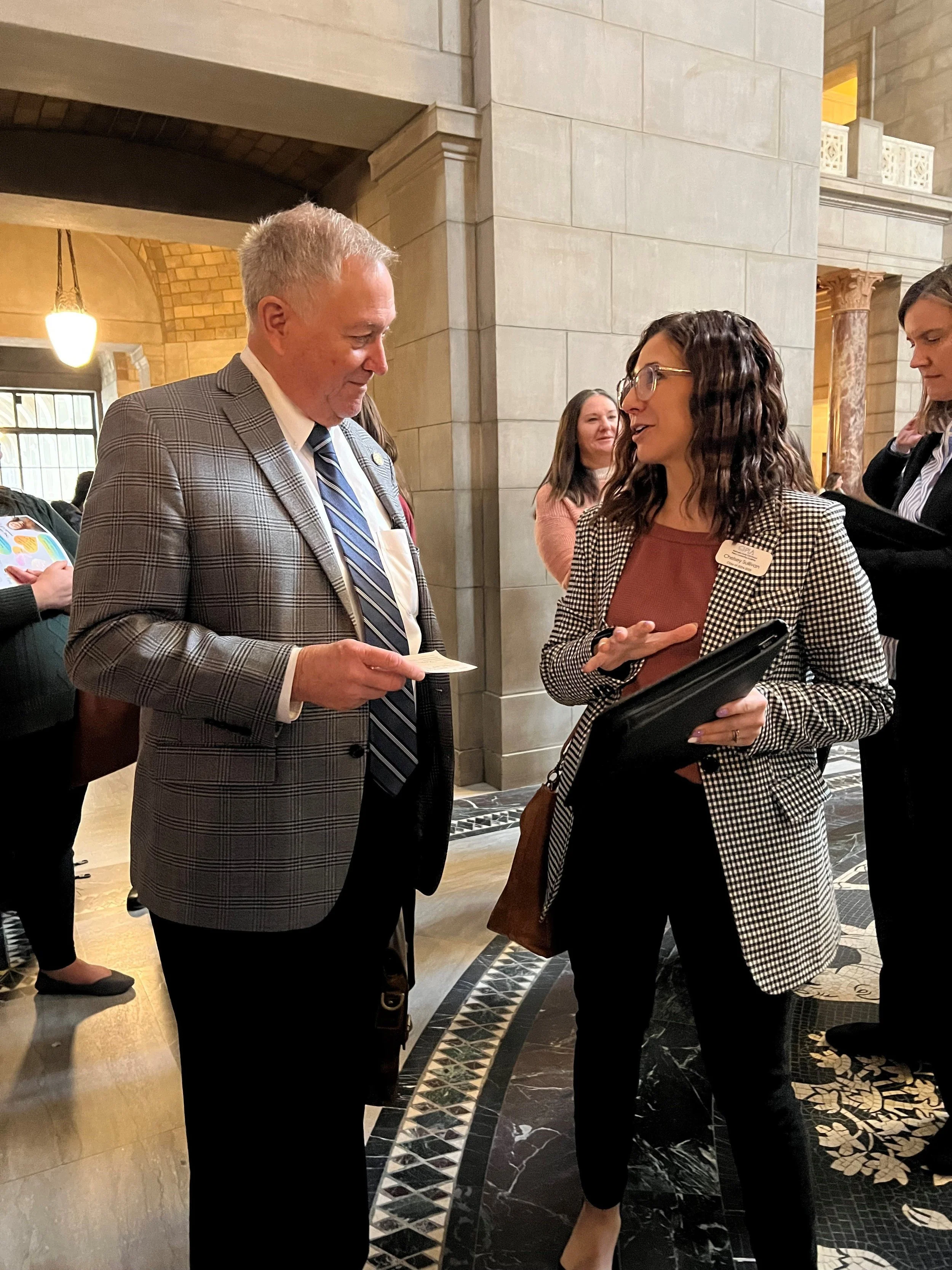 Woman with checkered jacket speaking with a man in a light gray suit jacket in the Rotunda of the Nebraska Capitol building.