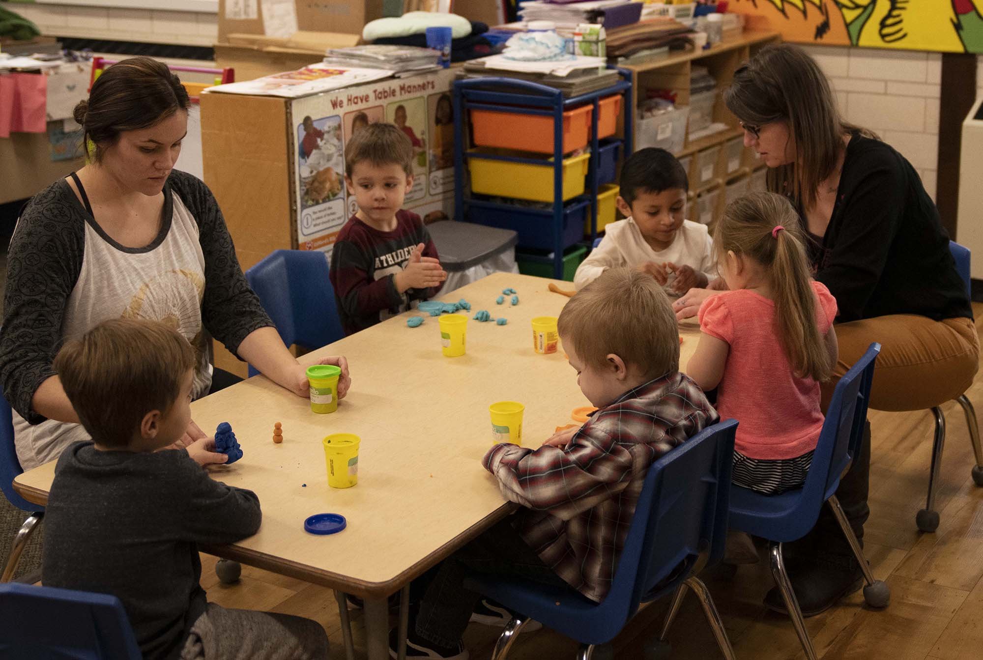 Two female teachers sit with a group of young children at an activity table in a child care center.