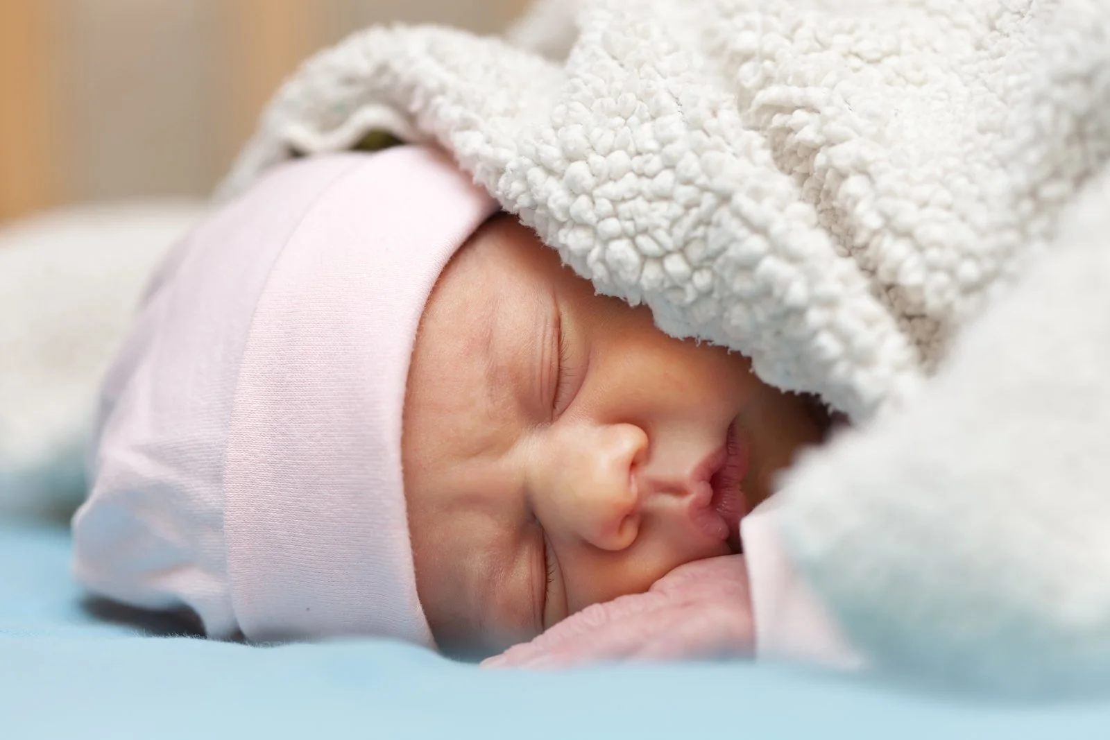 Newborn baby sleeping on its side, covered by a fuzzy blanket and wearing a pink fabric cap.