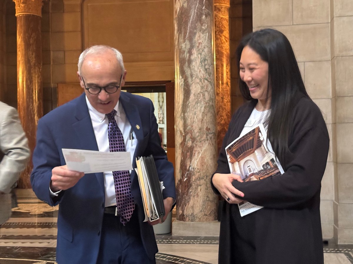 Woman in a dark jacket holding papers stands next to a man in a blue suit with glasses, carrying a binder and reading a handout.