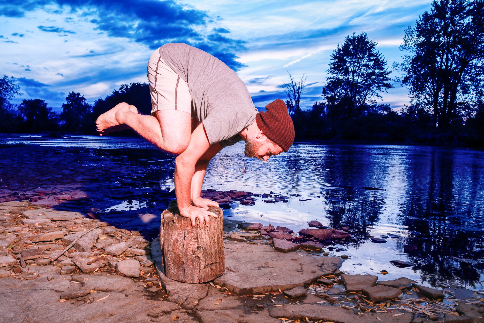 Man performing a handstand on a tree stump by a river during sunset.