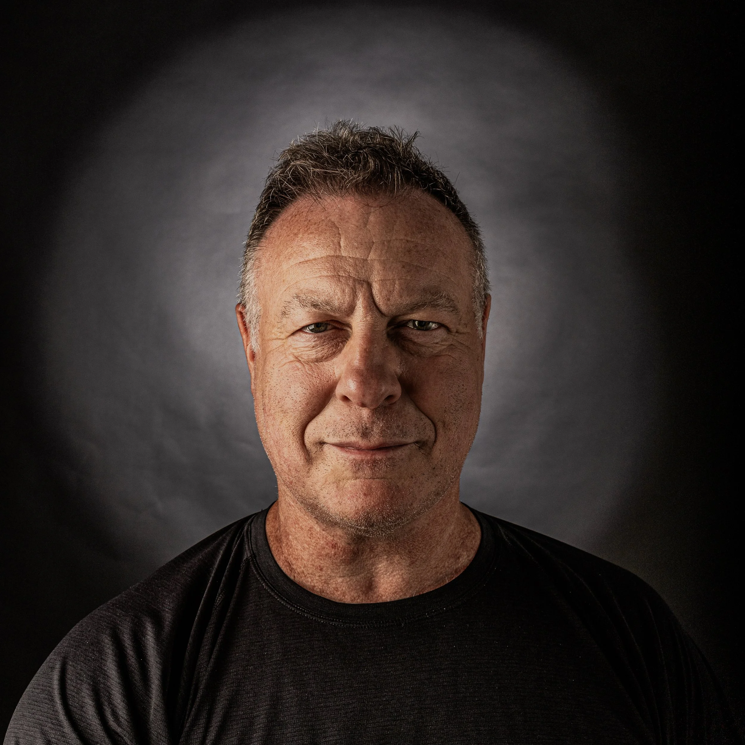 Close-up portrait of a middle-aged man with short, dark hair, wearing a black shirt, standing against a dark background with a subtle gradient.