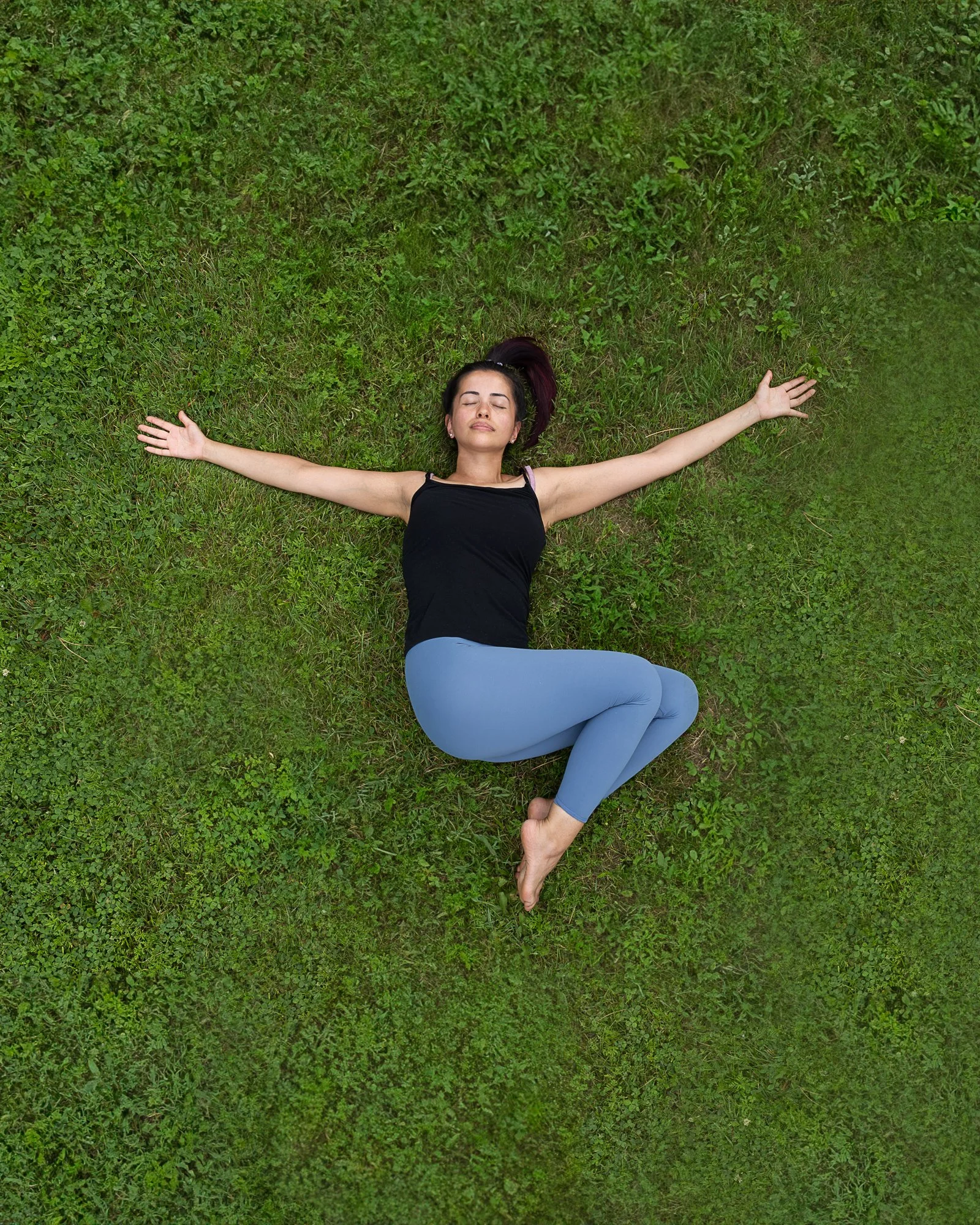 A woman lying on green grass with arms outstretched and eyes closed, practicing yoga or relaxation.