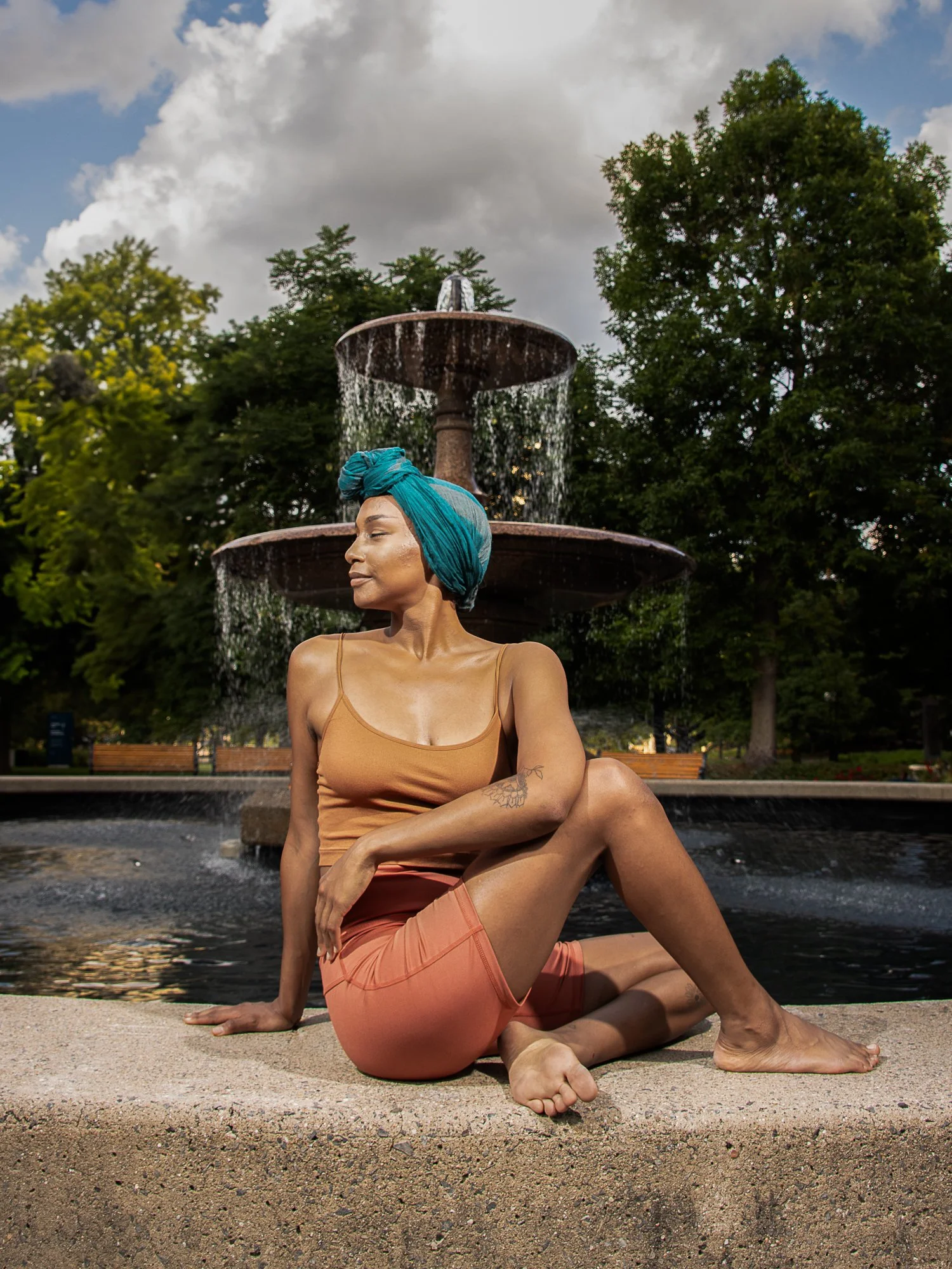 A woman sitting cross-legged on the edge of a fountain in a park, wearing a turquoise headwrap, a tan tank top, and peach shorts, surrounded by green trees and a cloudy sky.