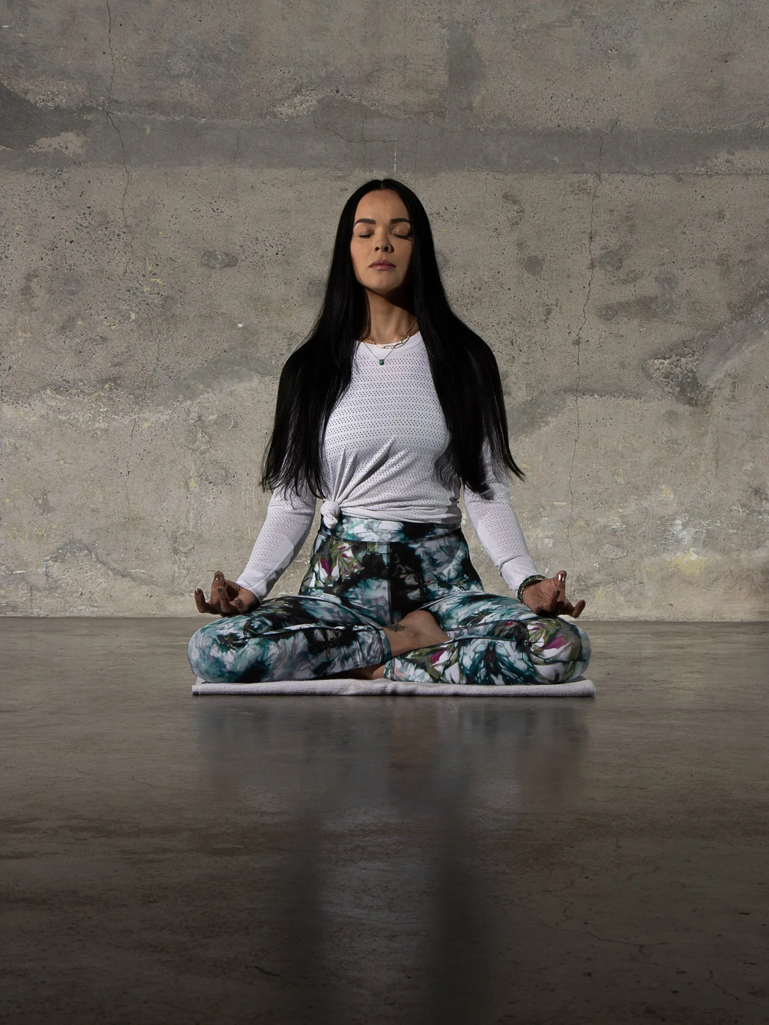 A woman practicing yoga in a cross-legged seated meditation pose on a mat in front of a concrete wall, with her eyes closed and hands resting on her knees.