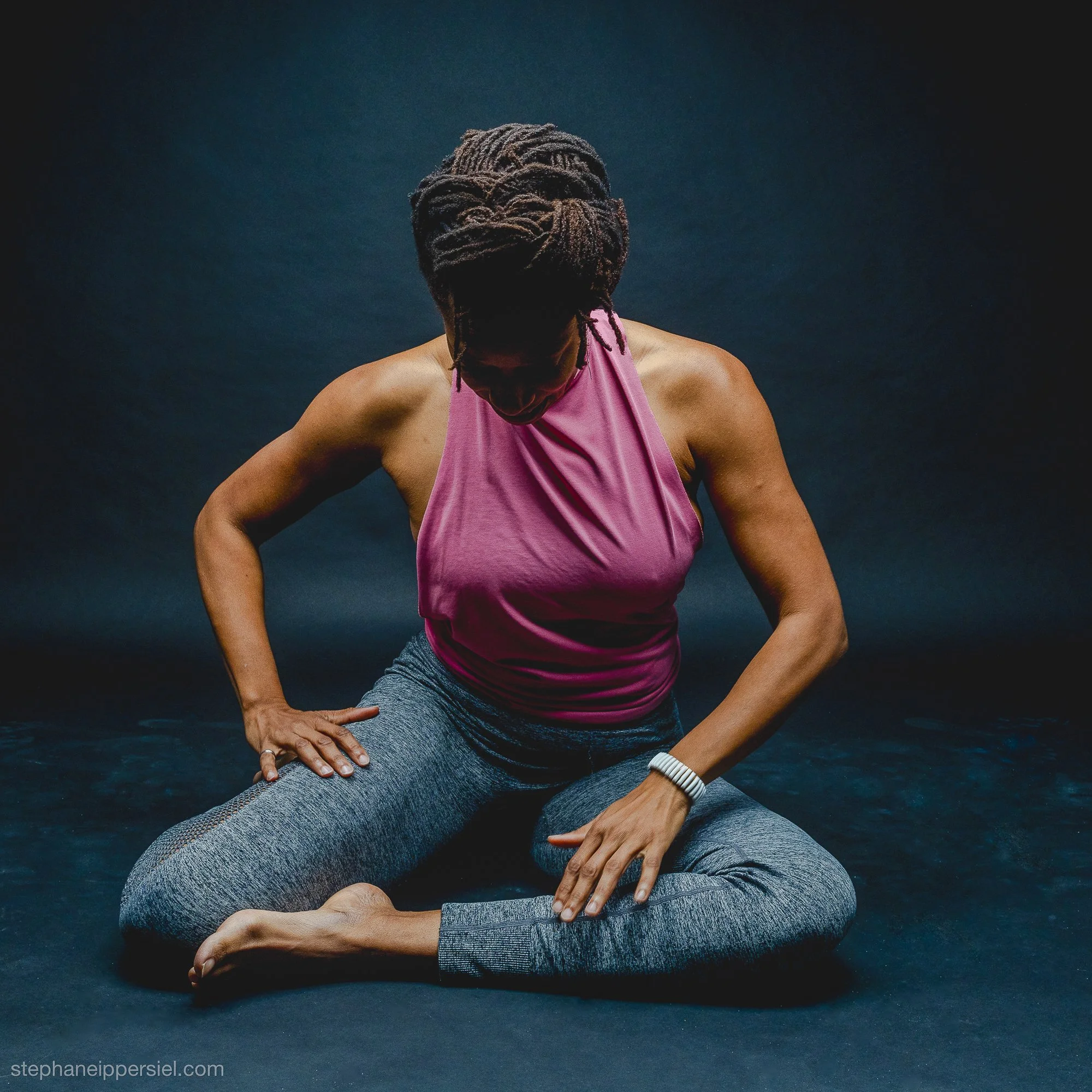 A woman practicing yoga in a kneeling pose, wearing a pink top and gray leggings, on a dark mat with a dark background.
