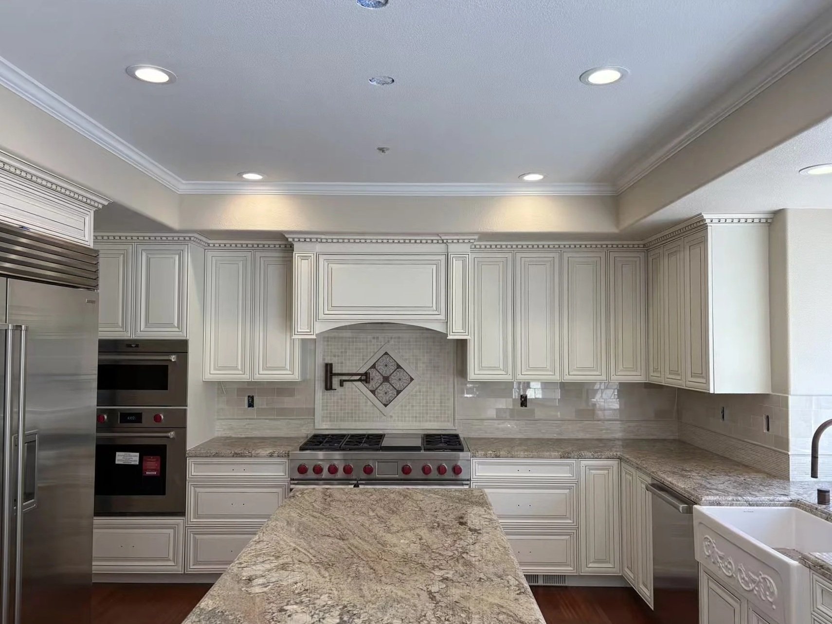 Kitchen with white cabinets, granite countertop island, stainless steel appliances, and beige tile backsplash, under recessed ceiling lights.