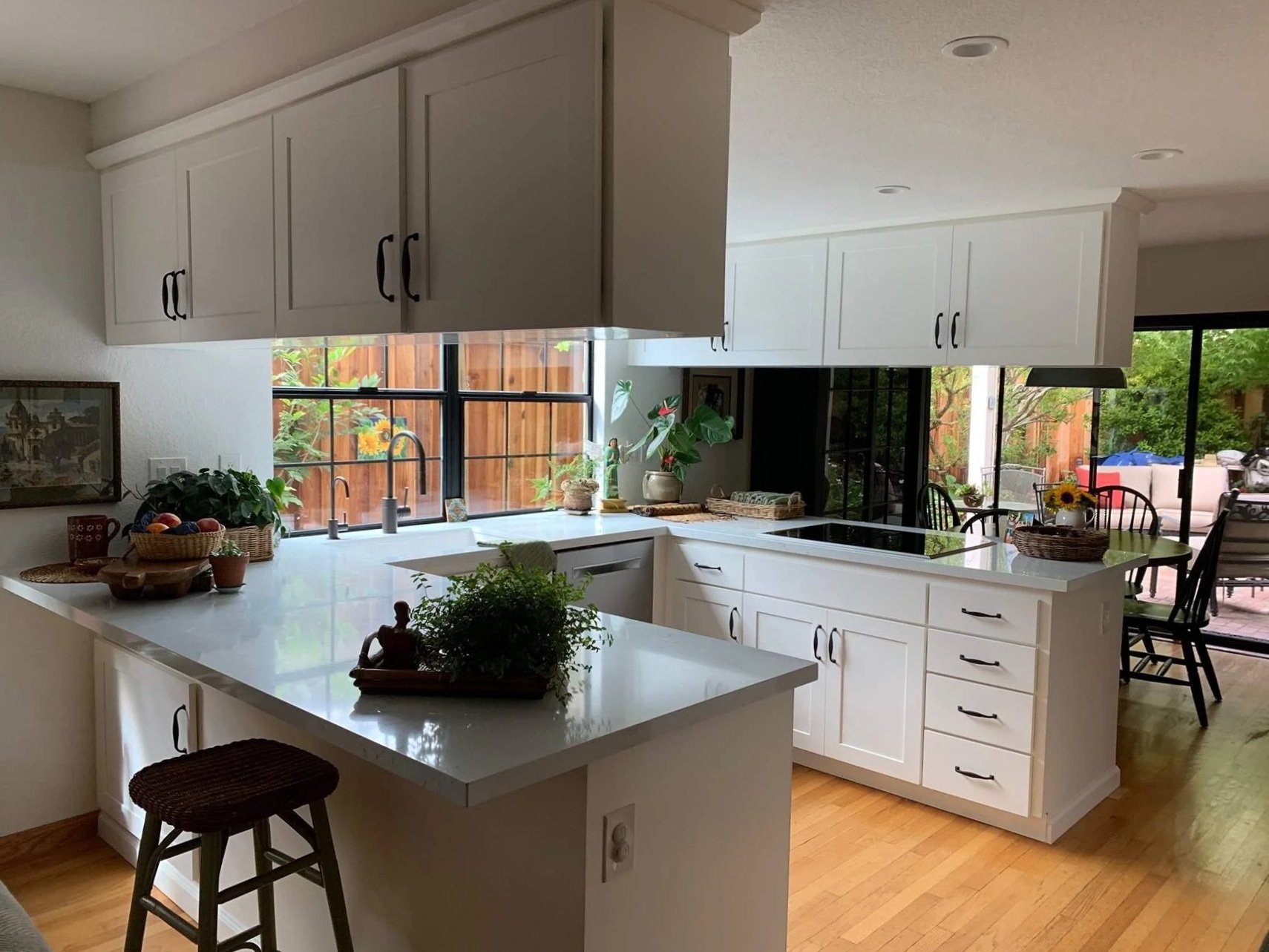 Bright kitchen with white cabinets, a white island with a dark stool, and a window with a view of a backyard with outdoor seating and green trees.
