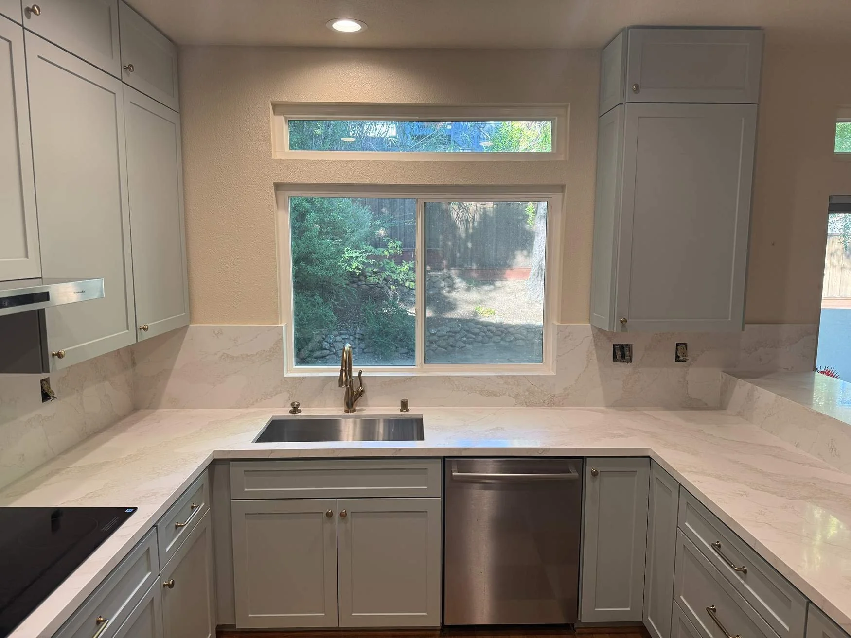 Kitchen with white countertops, white cabinets, a stainless steel sink, and a window overlooking a backyard with trees.