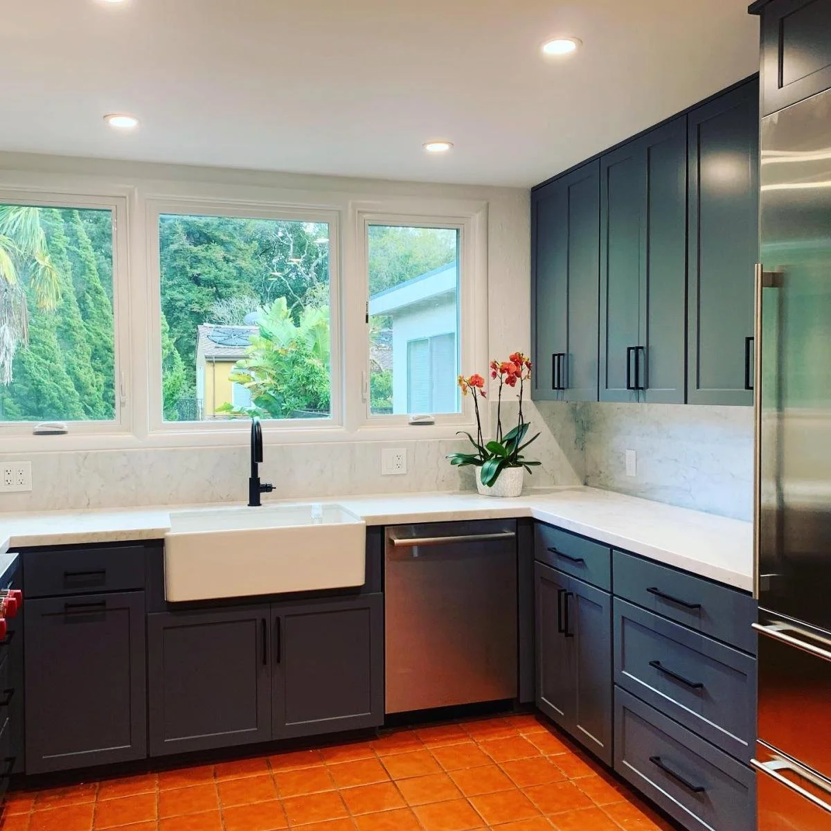 Modern kitchen with dark gray cabinets, white farmhouse sink, black faucet, stainless steel dishwasher, large window with green outdoor view, white marble countertops, potted orange orchid, terracotta tile floor.