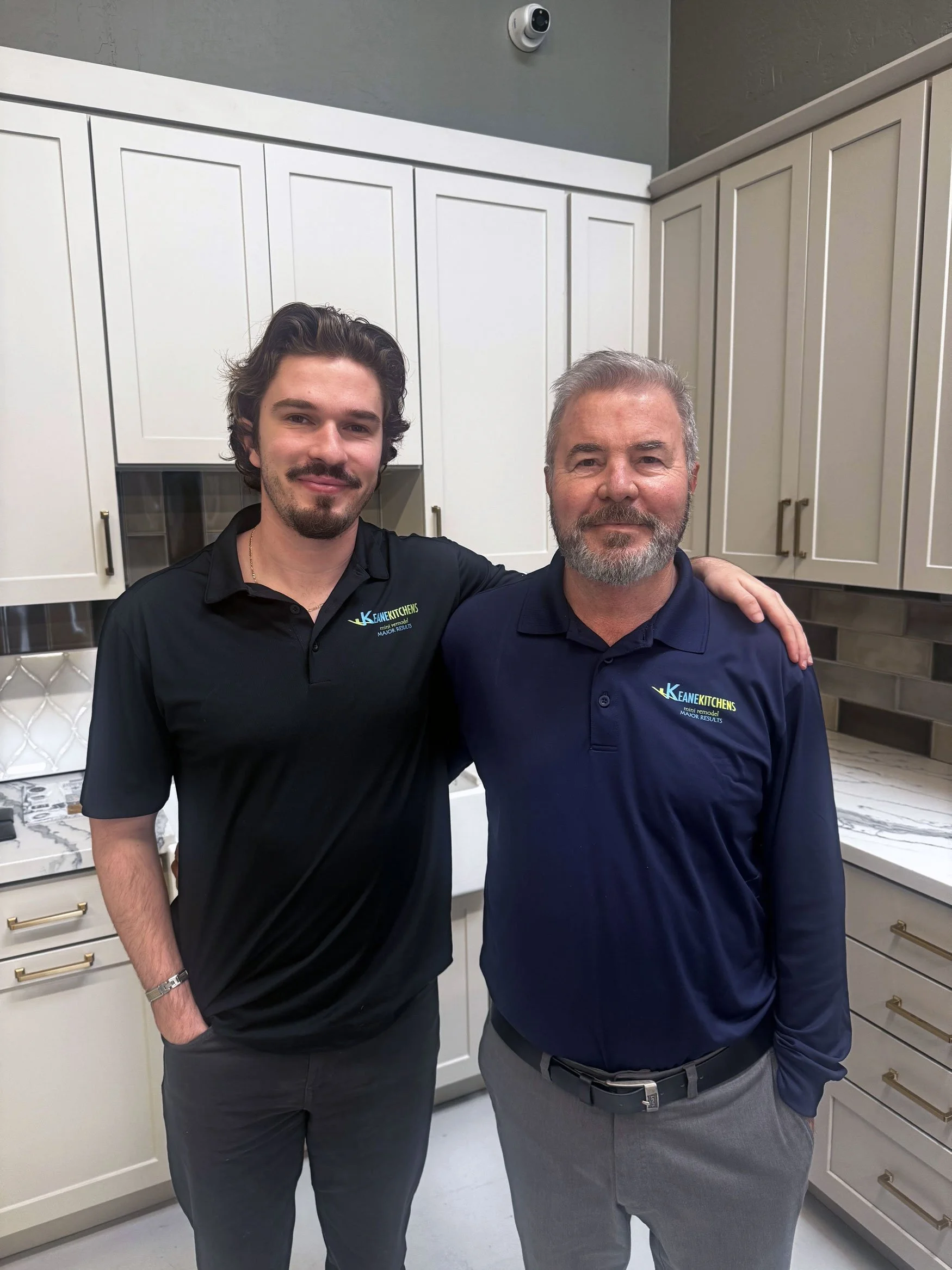 Two men standing in a kitchen with white cabinets, wearing matching navy blue shirts with 'Keane Kitchens' logo, smiling with one man's arm around the other's shoulder.