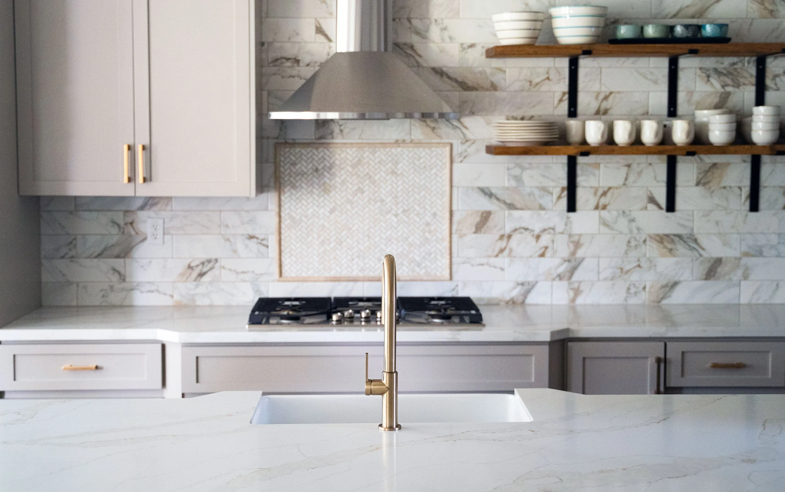 Modern kitchen with marble backsplash, white countertops, gold cabinet handles, open wooden shelves with bowls and cups, and a gold faucet at the sink.