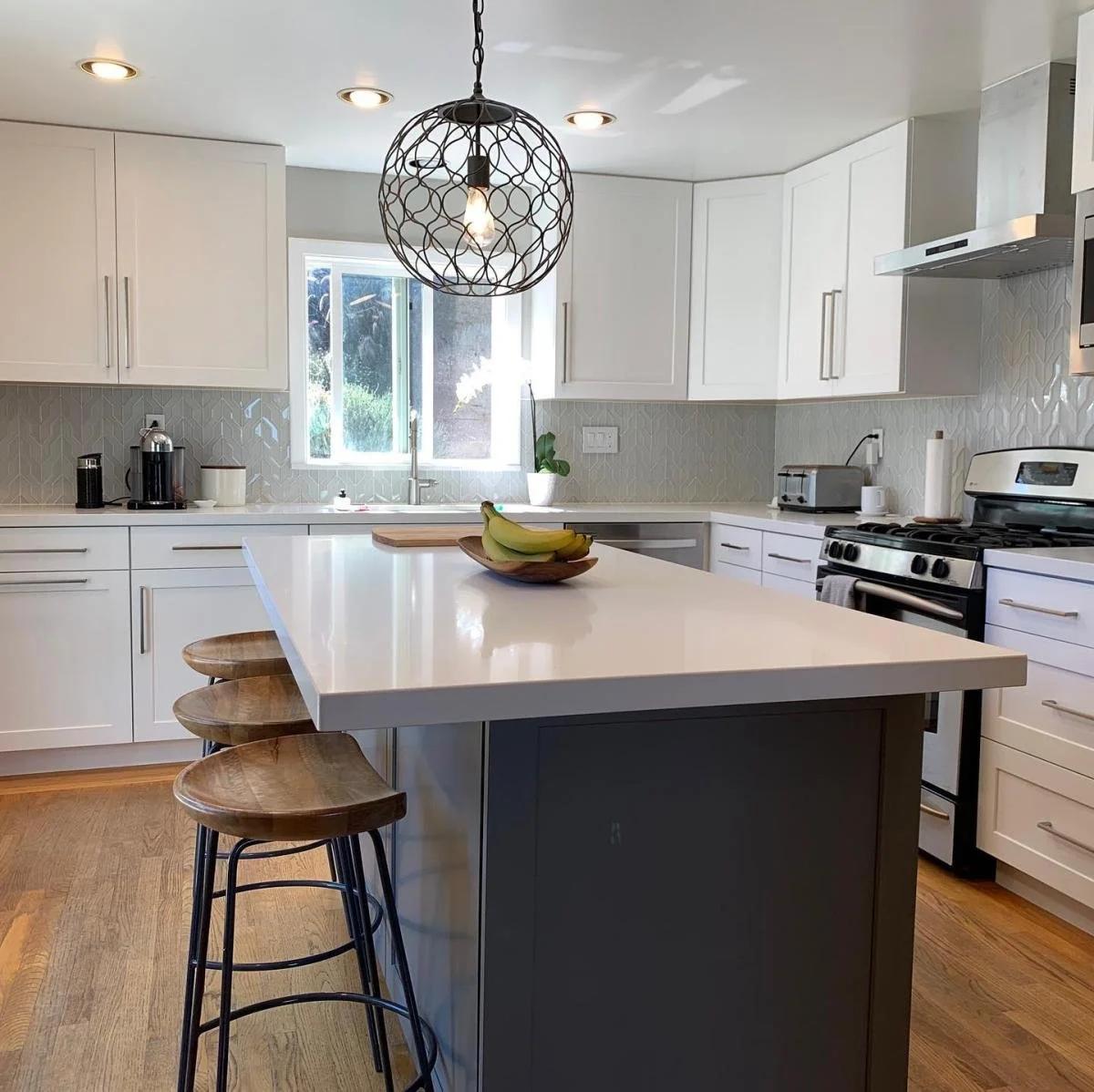 Modern kitchen with white cabinets, a central island with a white countertop, a bowl of bananas, three wooden stools with metal legs, a pendant light, a window, and various appliances including a stove, toaster, and coffee maker.