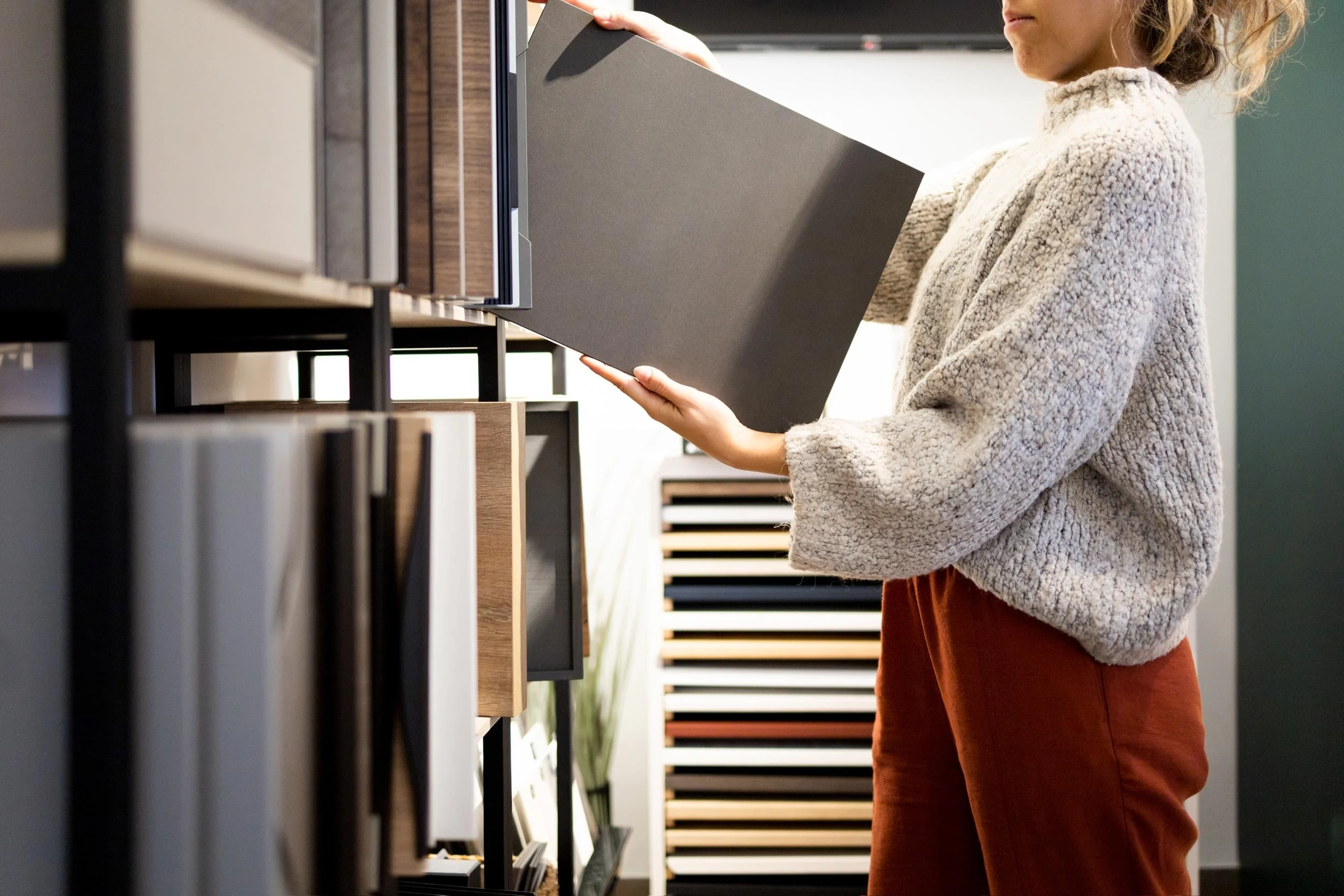 A woman options different styles of laminate flooring samples in a showroom, holding a sample in her left hand and examining another with her right hand.