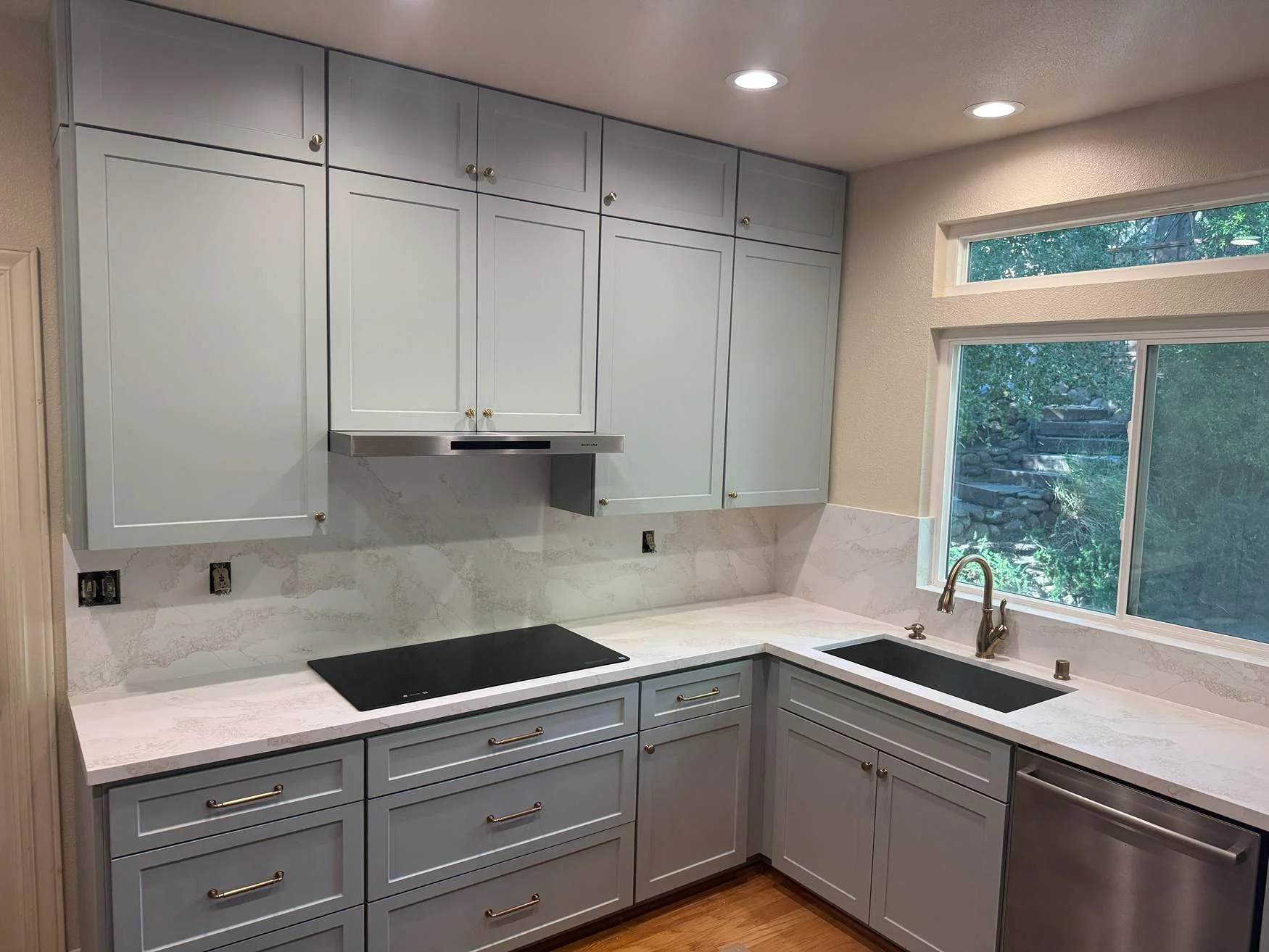 Modern kitchen with white cabinets, marble backsplash, black stovetop, stainless steel range hood, black sink, and large window overlooking greenery.