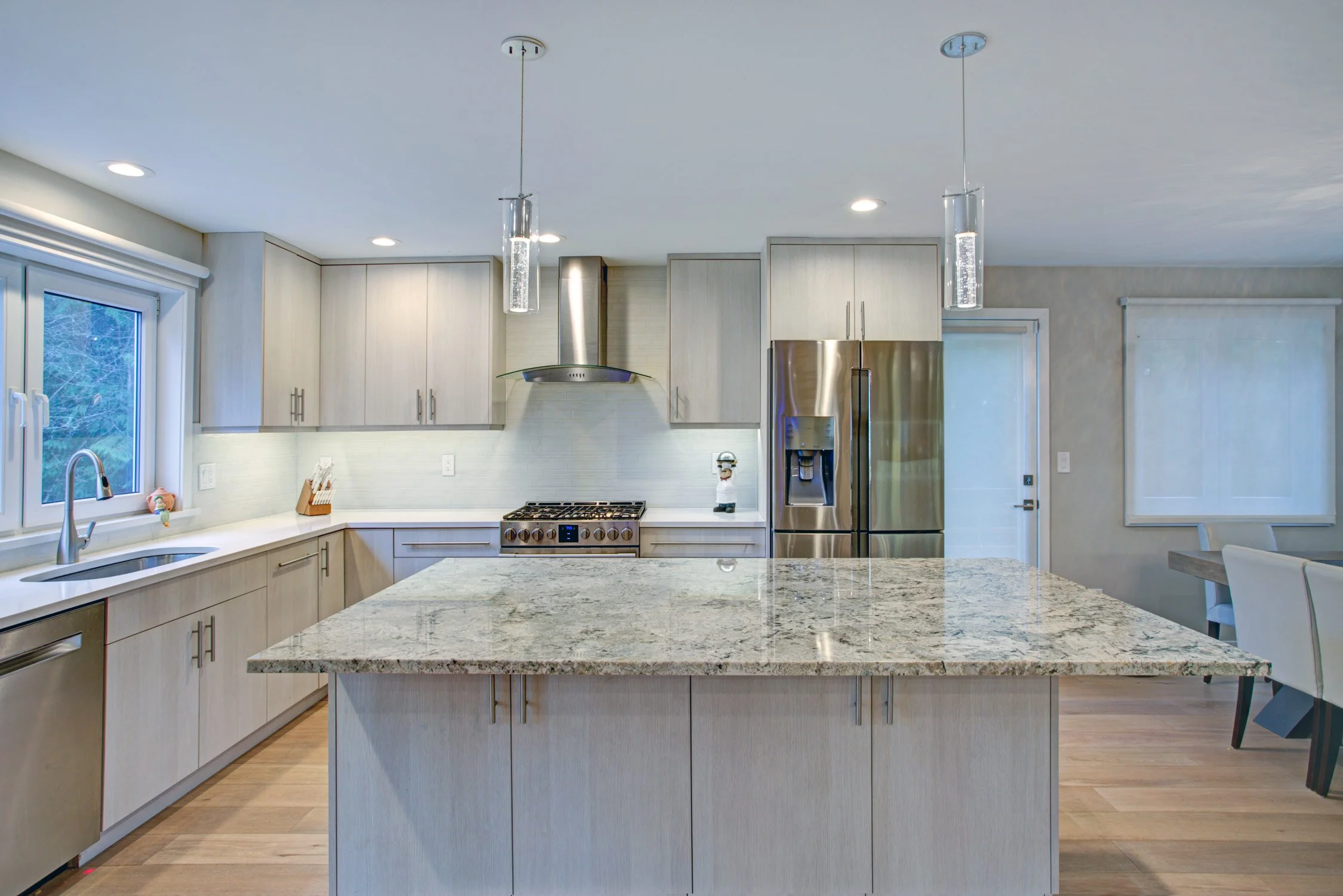 Modern kitchen with light wood cabinets, granite island, stainless steel appliances, pendant lighting, and a window above the sink.