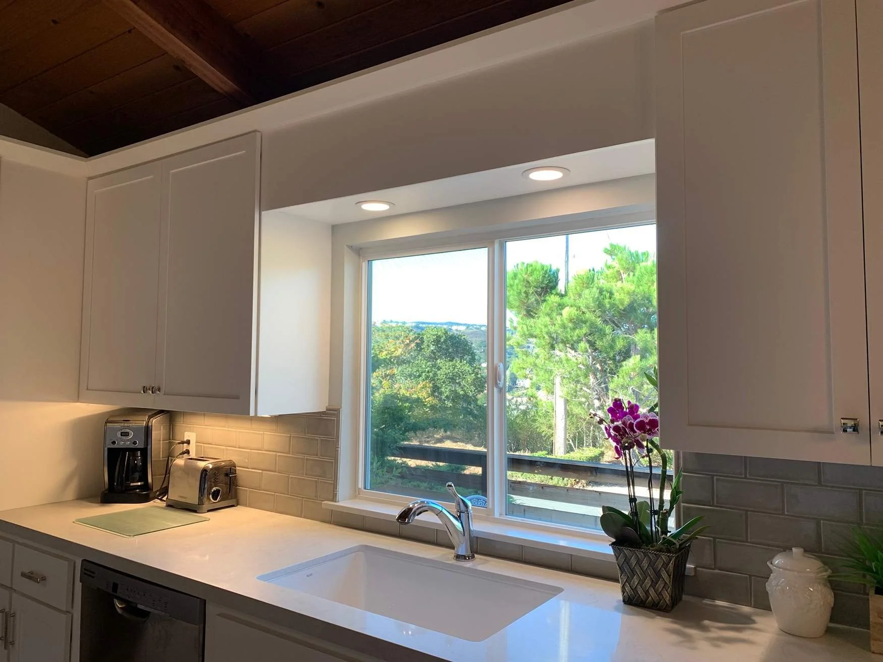 A kitchen with white cabinets, a white countertop, a window overlooking green trees, purple orchids, a coffee maker, a toaster, a kettle, and a sink.