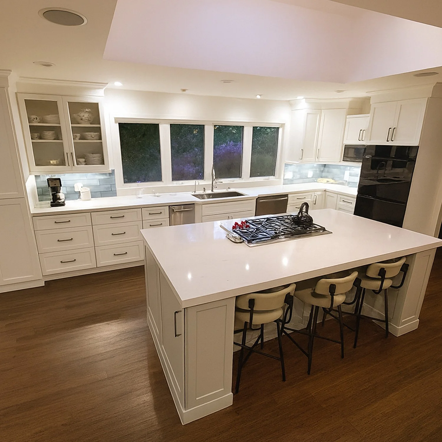 A modern kitchen with white cabinets, a large island with a white countertop, and three barstools. There are large windows above the sink, a stove on the island, and a black oven built into the cabinetry. The floor is wood, and the lighting is warm a