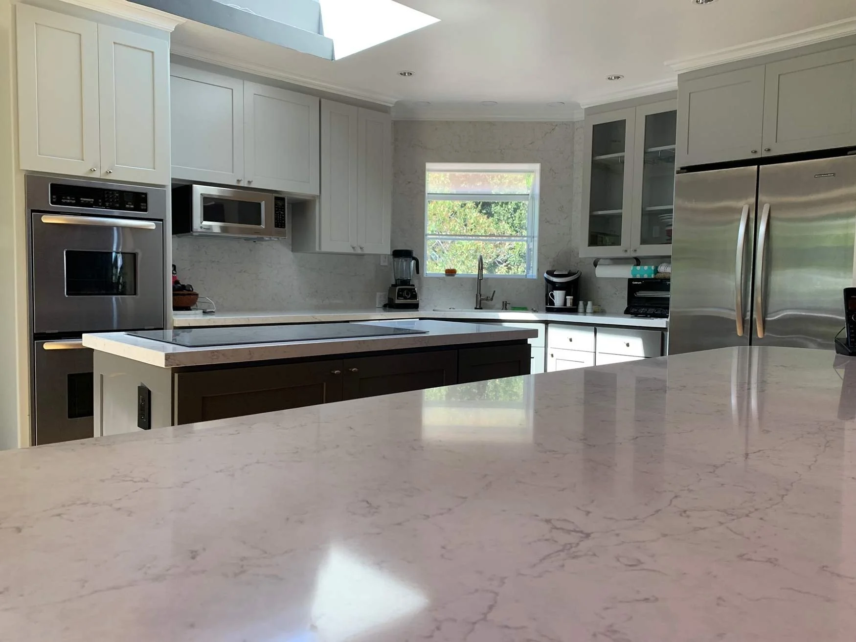 Modern kitchen with white and dark cabinetry, marble countertops, stainless steel appliances including a refrigerator, oven, microwave, and a stove top, with a window above the sink and various small appliances on the counters.