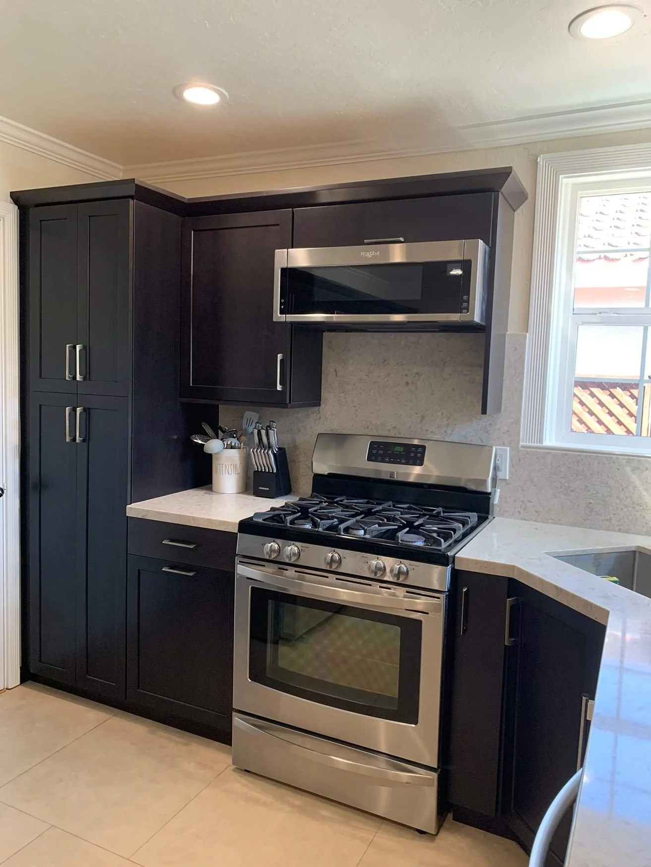 Kitchen with dark cabinets, stainless steel gas stove, microwave above, beige countertops, and a window with white trim.