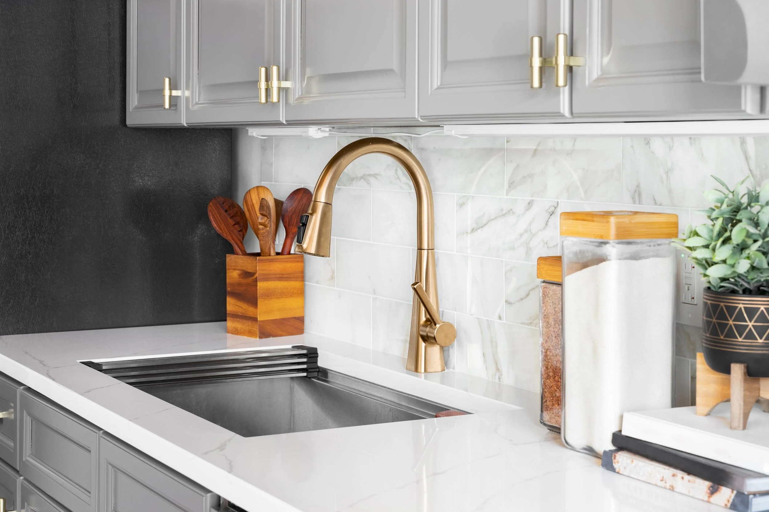 Modern kitchen with white marble backsplash, gold faucet, black sink, wooden utensils, and decorative containers.