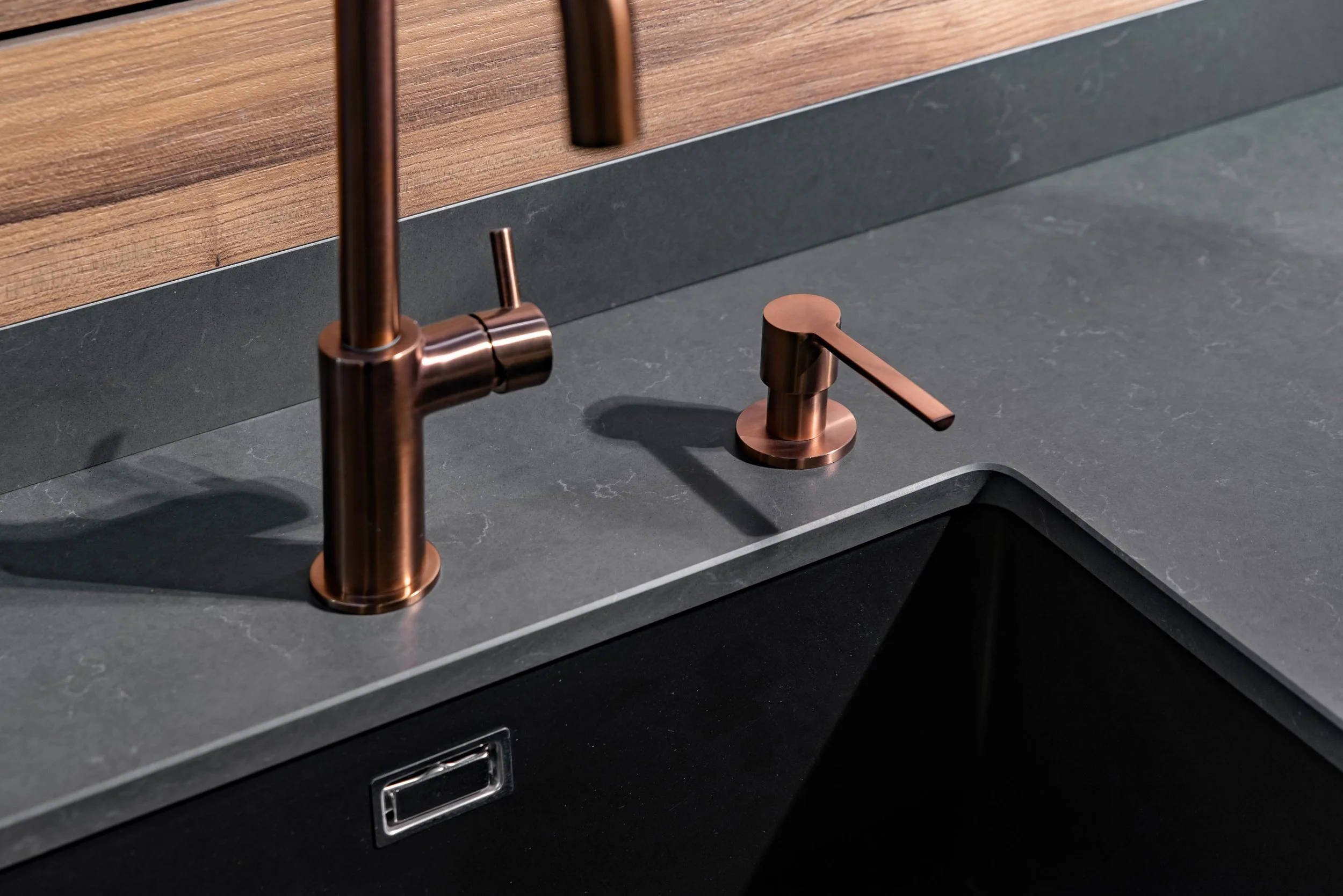 Close-up of a copper-colored kitchen faucet and soap dispenser on a dark gray countertop with a wooden backsplash.