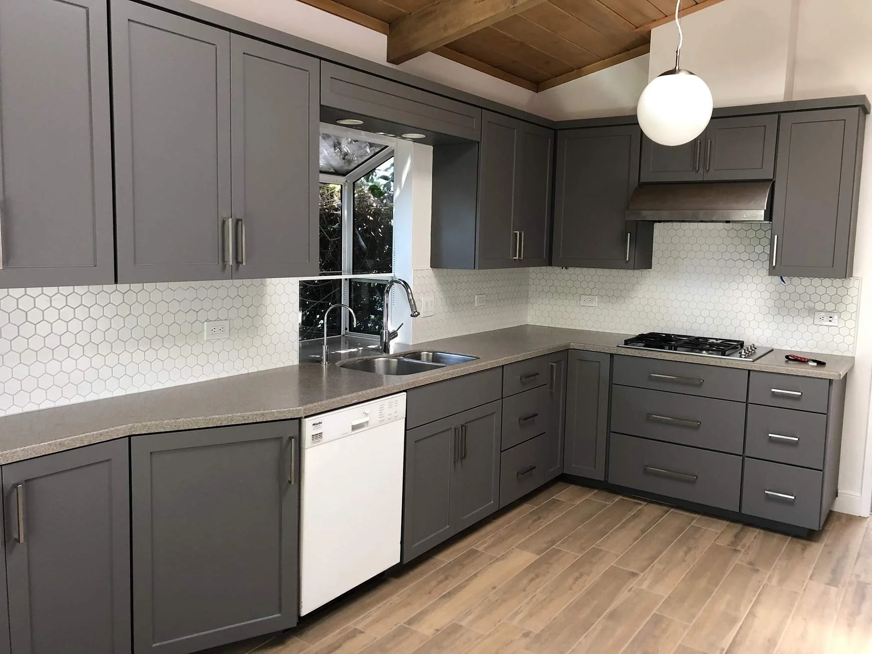 Modern kitchen with gray cabinets, white hexagonal tile backsplash, gray countertop, and wooden flooring. Includes a stainless steel sink, dishwasher, gas stove, and a round white pendant light.