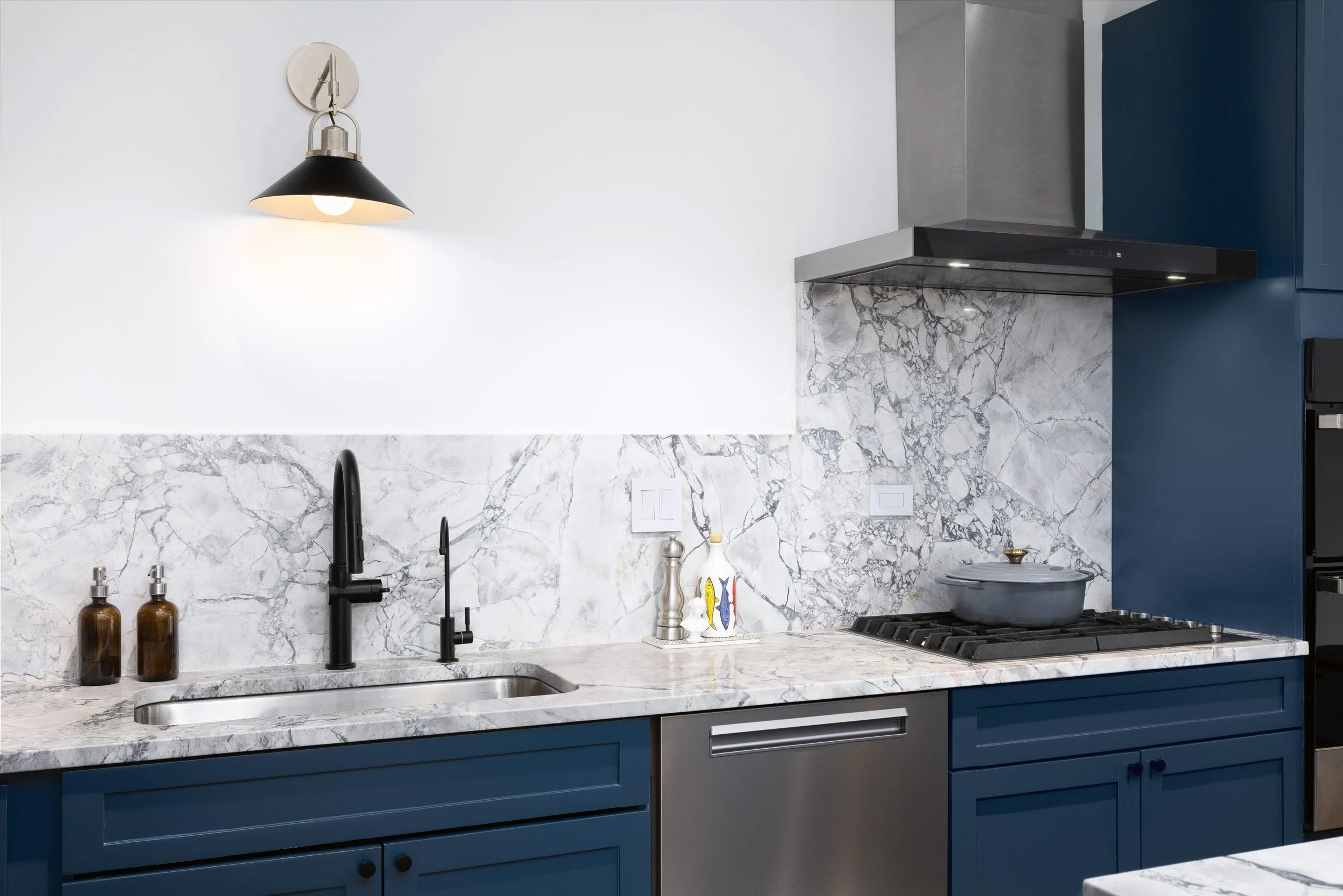 Modern kitchen with navy blue cabinets, marble countertops, white marble backsplash, black faucet, and wall-mounted light fixture.