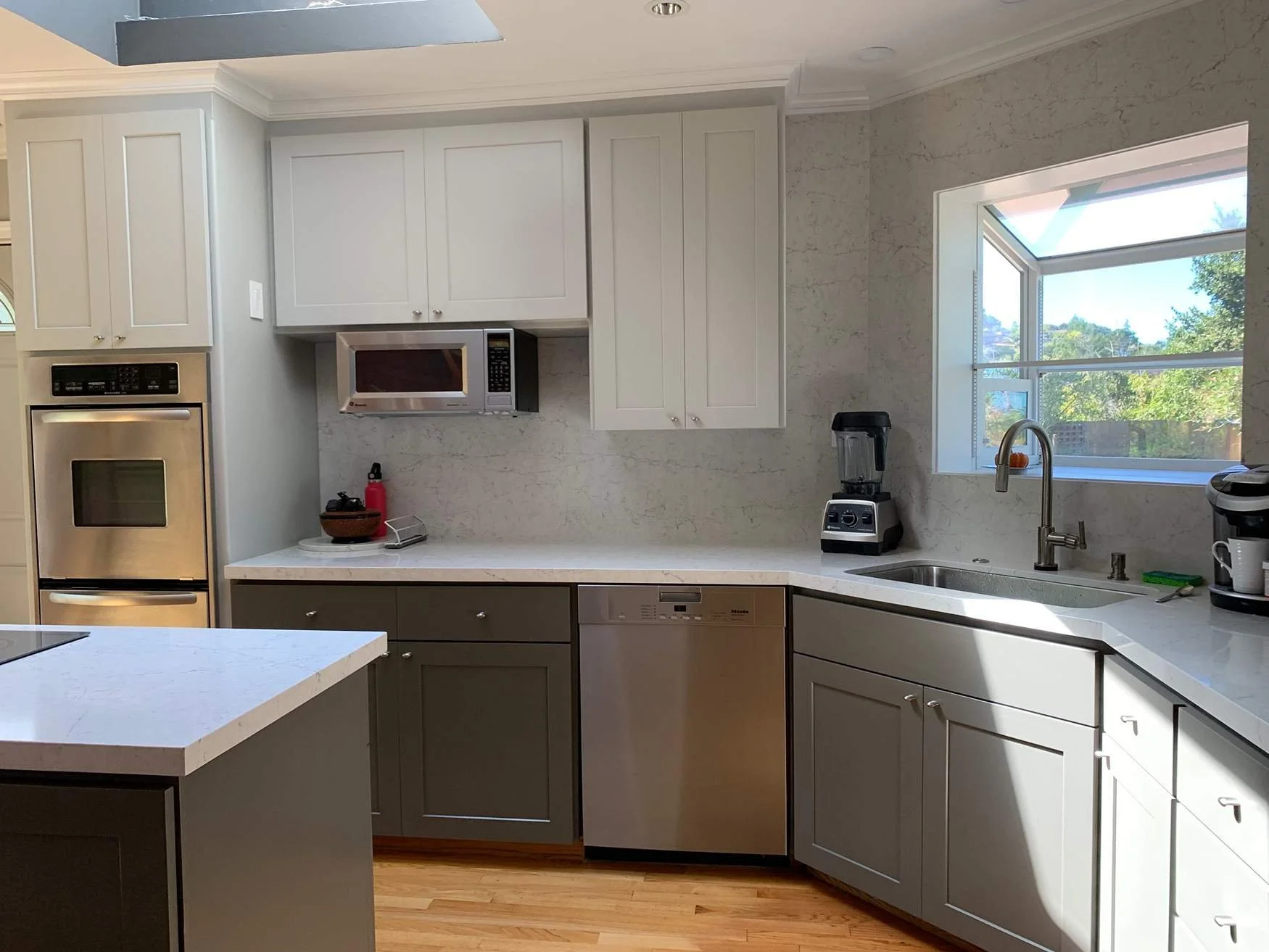Kitchen with white and gray cabinets, a window, stainless steel microwave, oven, and dishwasher, with small appliances on the counter.