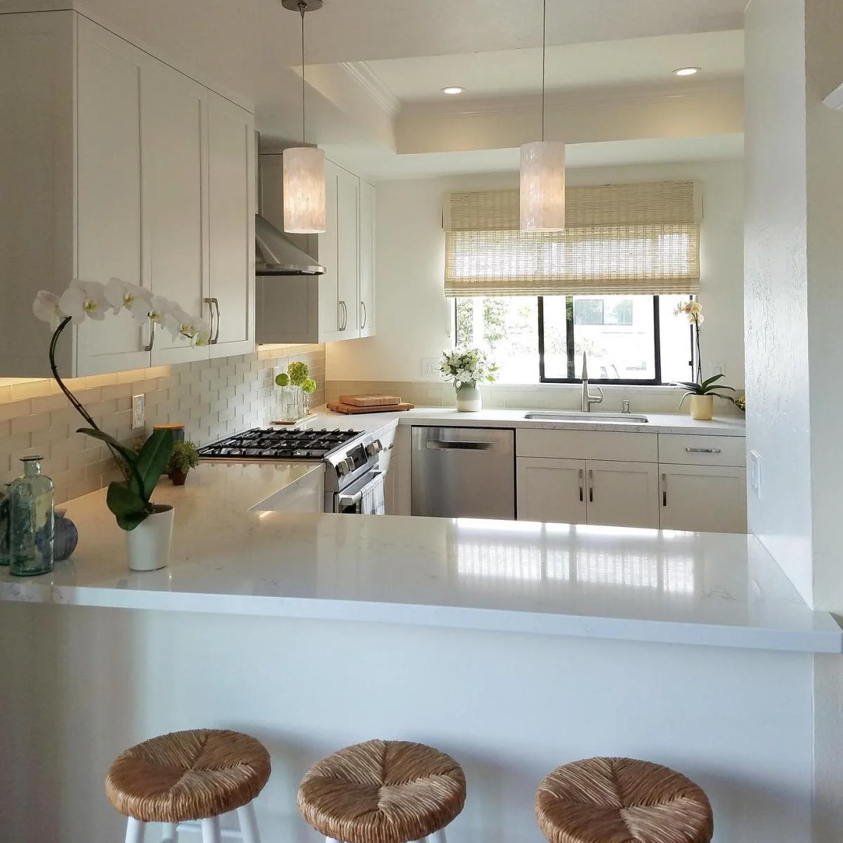 A bright, modern kitchen with white cabinets, a stove, and stainless steel dishwasher. Decor includes plants and flowers on the counter and windowsill, with a window above the sink. There are three wicker stools at the counter.