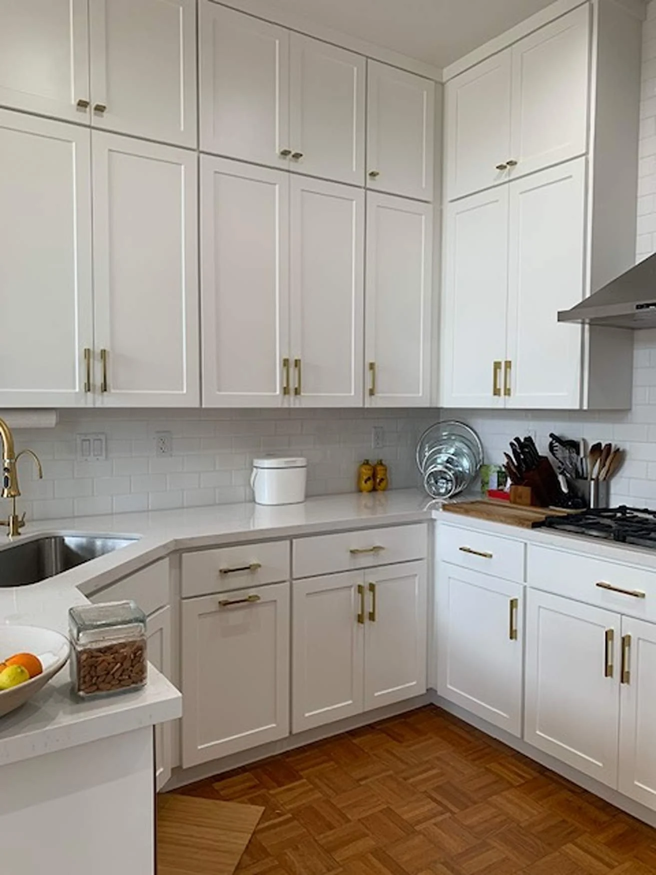 A clean, white kitchen with white cabinets with gold handles, a white countertop, and a wood floor. There are various kitchen items including a bowl of fruit, a jar of almonds, dish racks with dishes, and kitchen utensils.