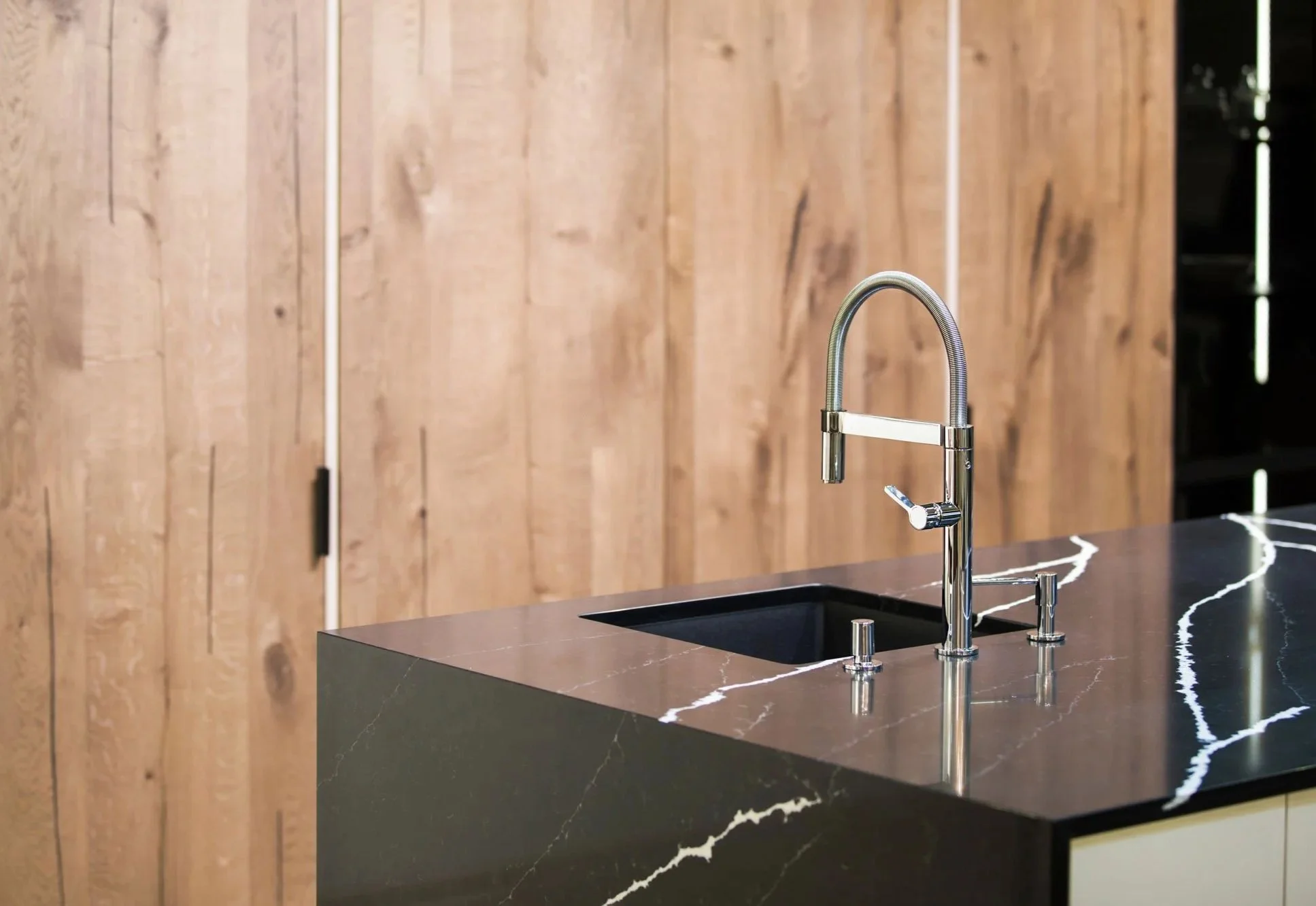 Modern kitchen island with black marble countertop and a sleek chrome faucet, wooden wall in the background.
