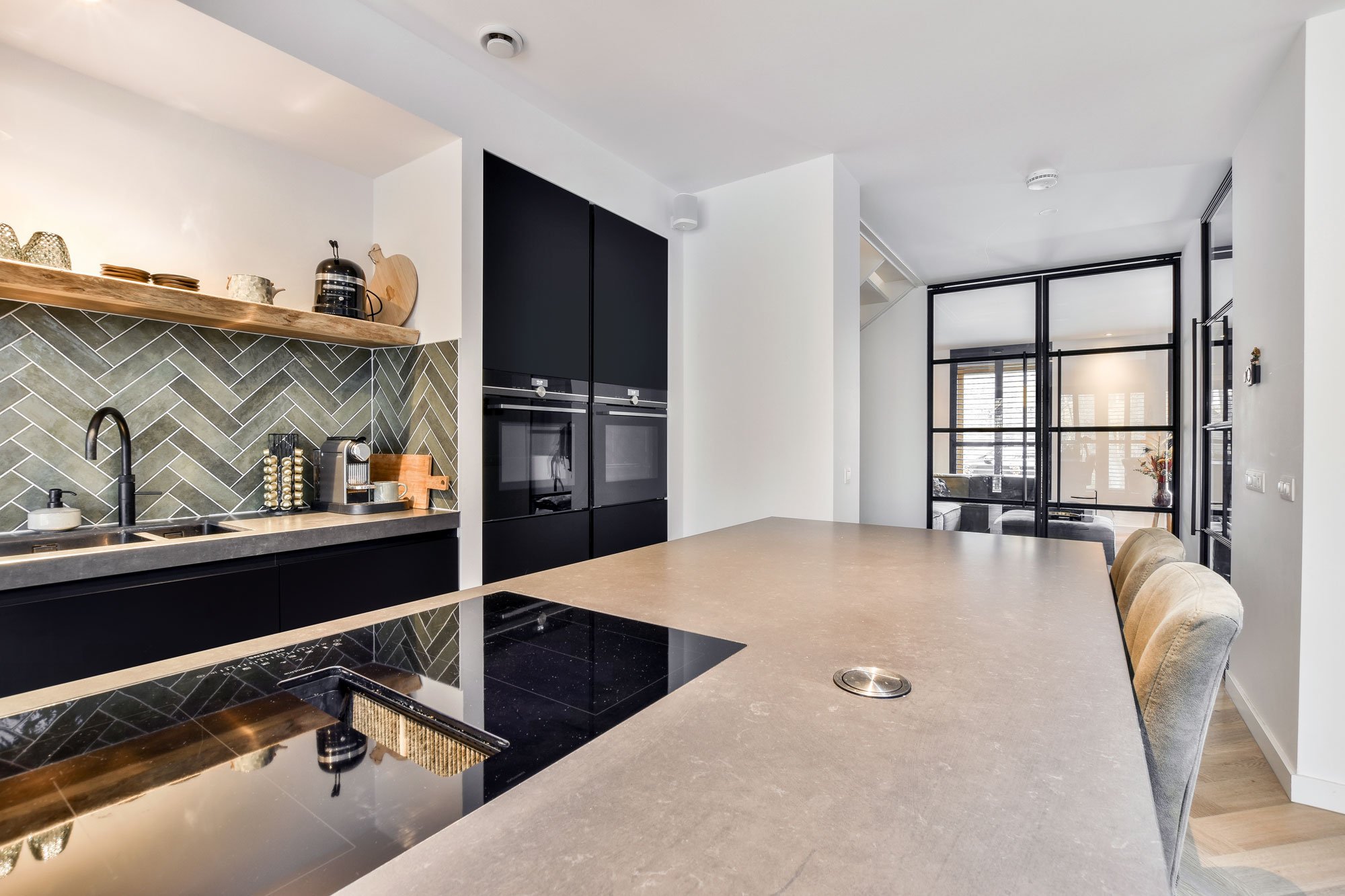 Modern kitchen with black cabinets, a beige countertop, green herringbone backsplash, and barstools at the counter.