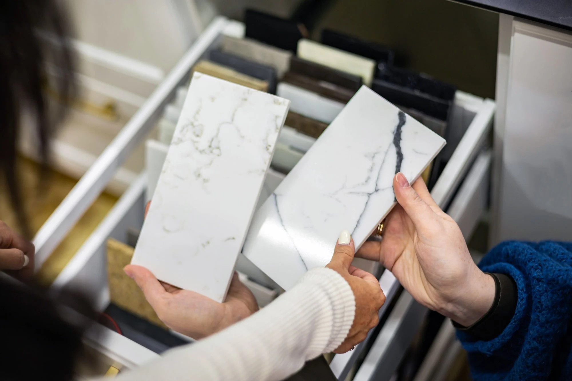 Two people are holding marble-look tile samples, with the background showing a display rack of various tile samples.