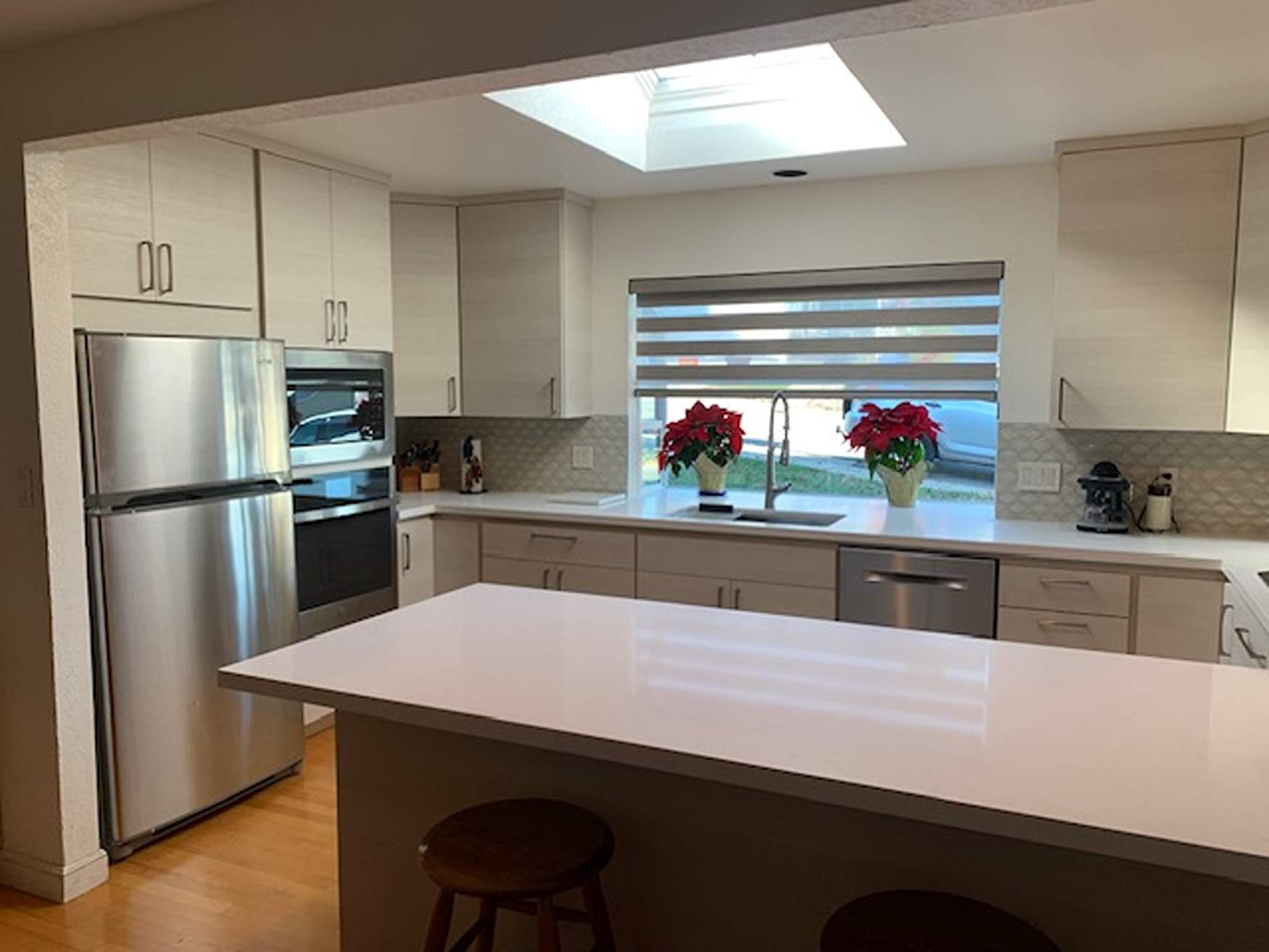 Modern kitchen with beige cabinets, stainless steel refrigerator and oven, a large white island, a window with striped blinds, and decorative red poinsettia plants.